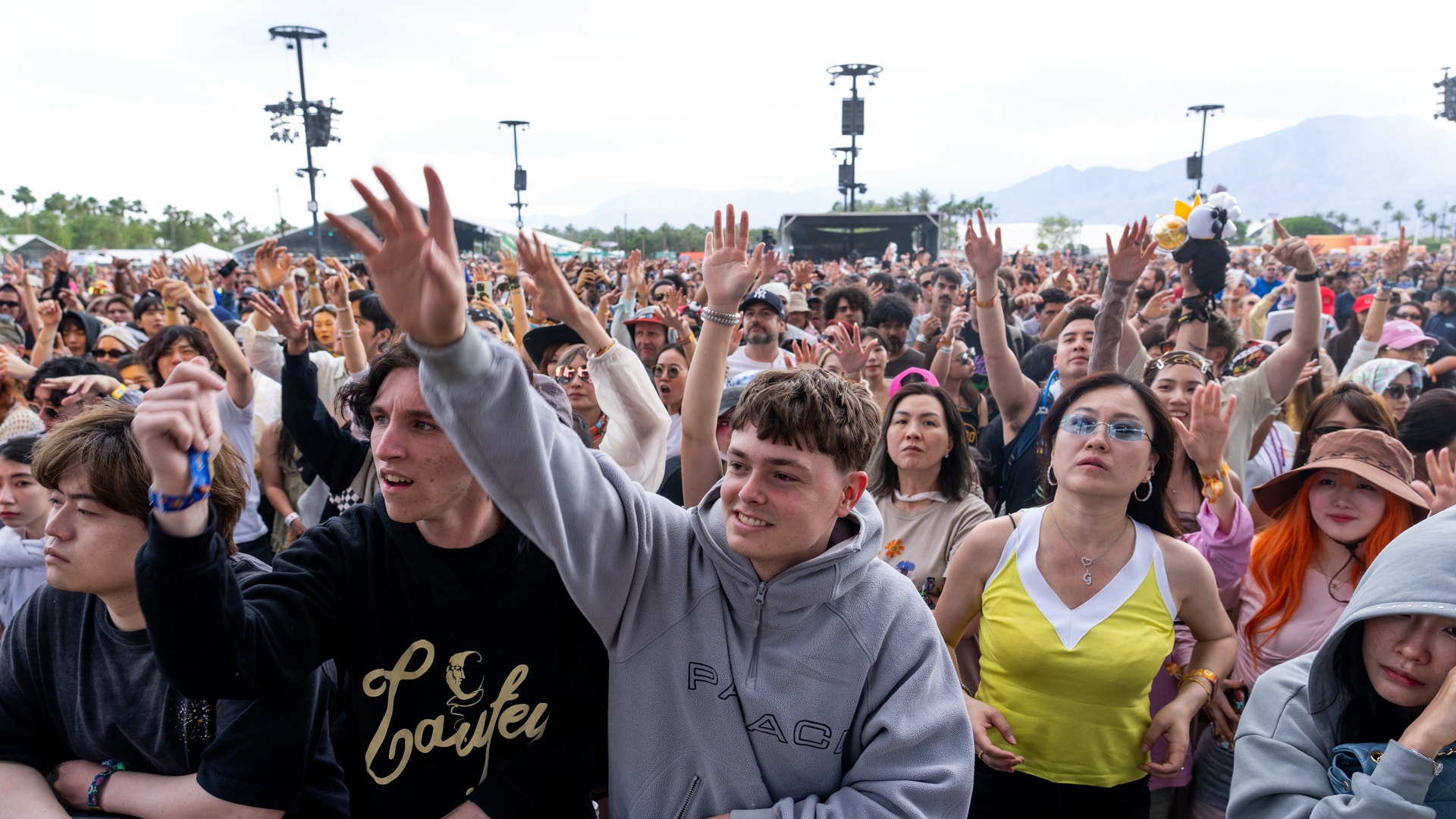 En stor folksamling på en musikfestival. Många människor står tätt tillsammans med upphöjda händer och njuter av konserten. Scenutrustning och berg syns i bakgrunden.
