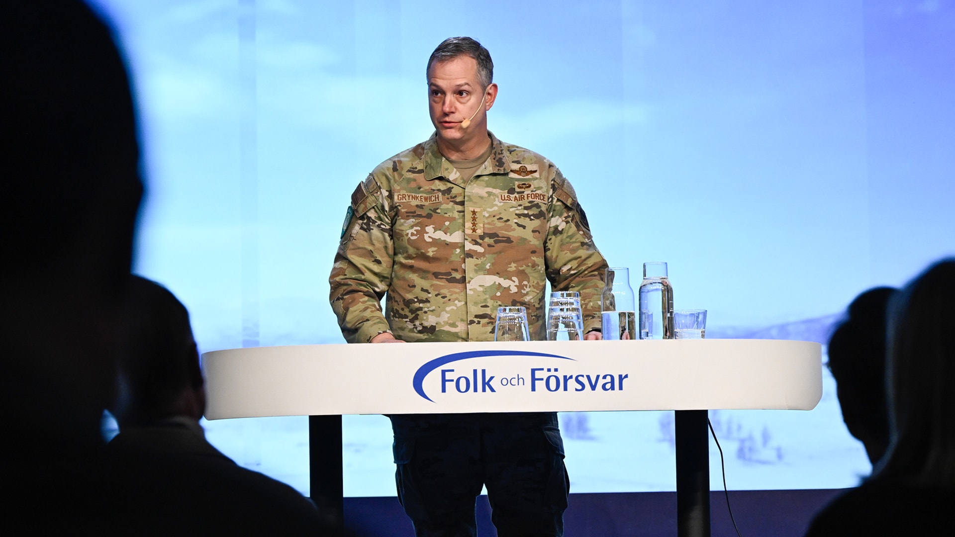 En man i militär uniform står bakom ett podium märkt Folk och Försvar och håller ett tal på scenen.