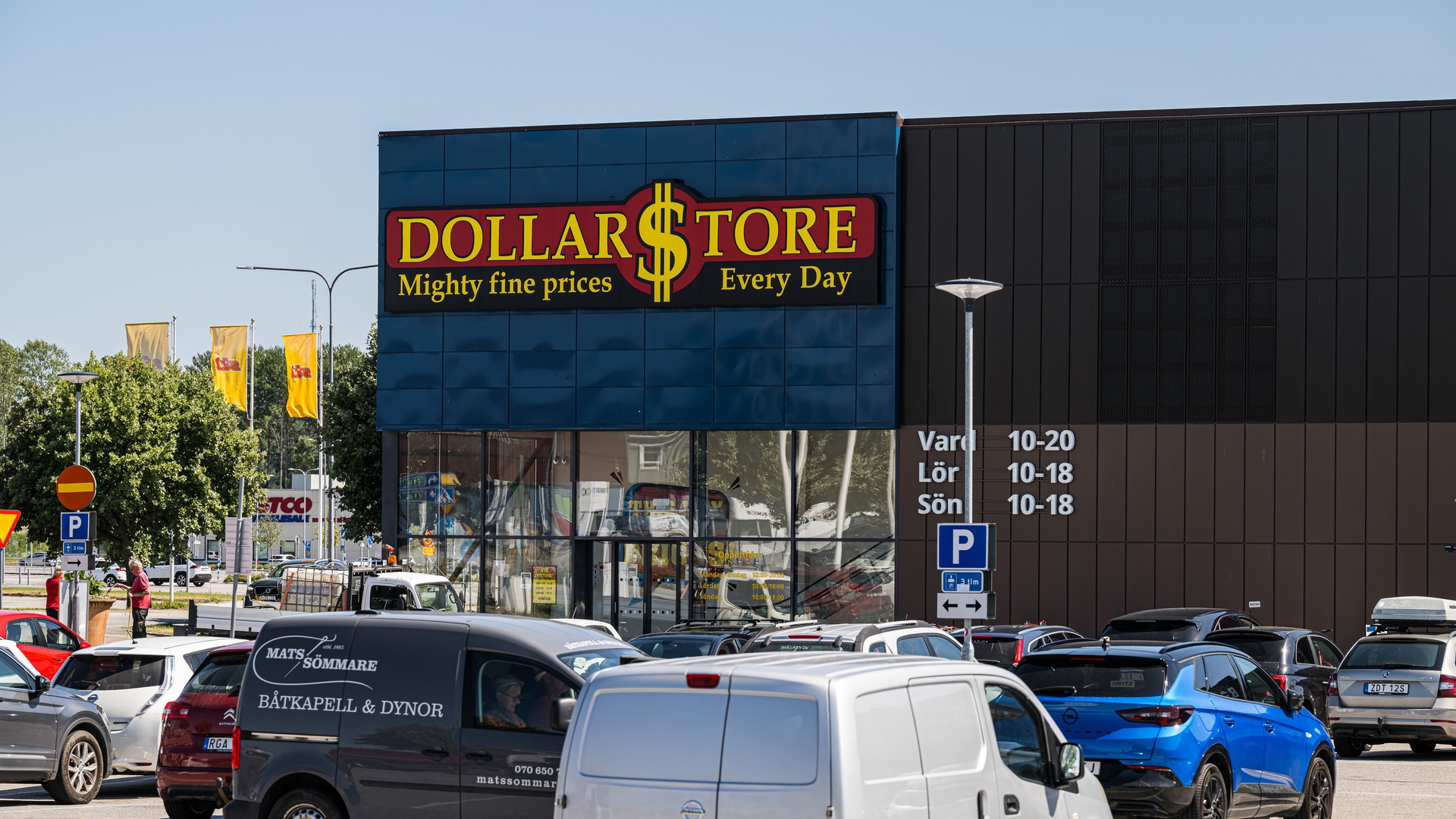 En Dollar Store-butik med blå fasad och gul logotyp. Framför butiken ligger en parkering med parkerade bilar.