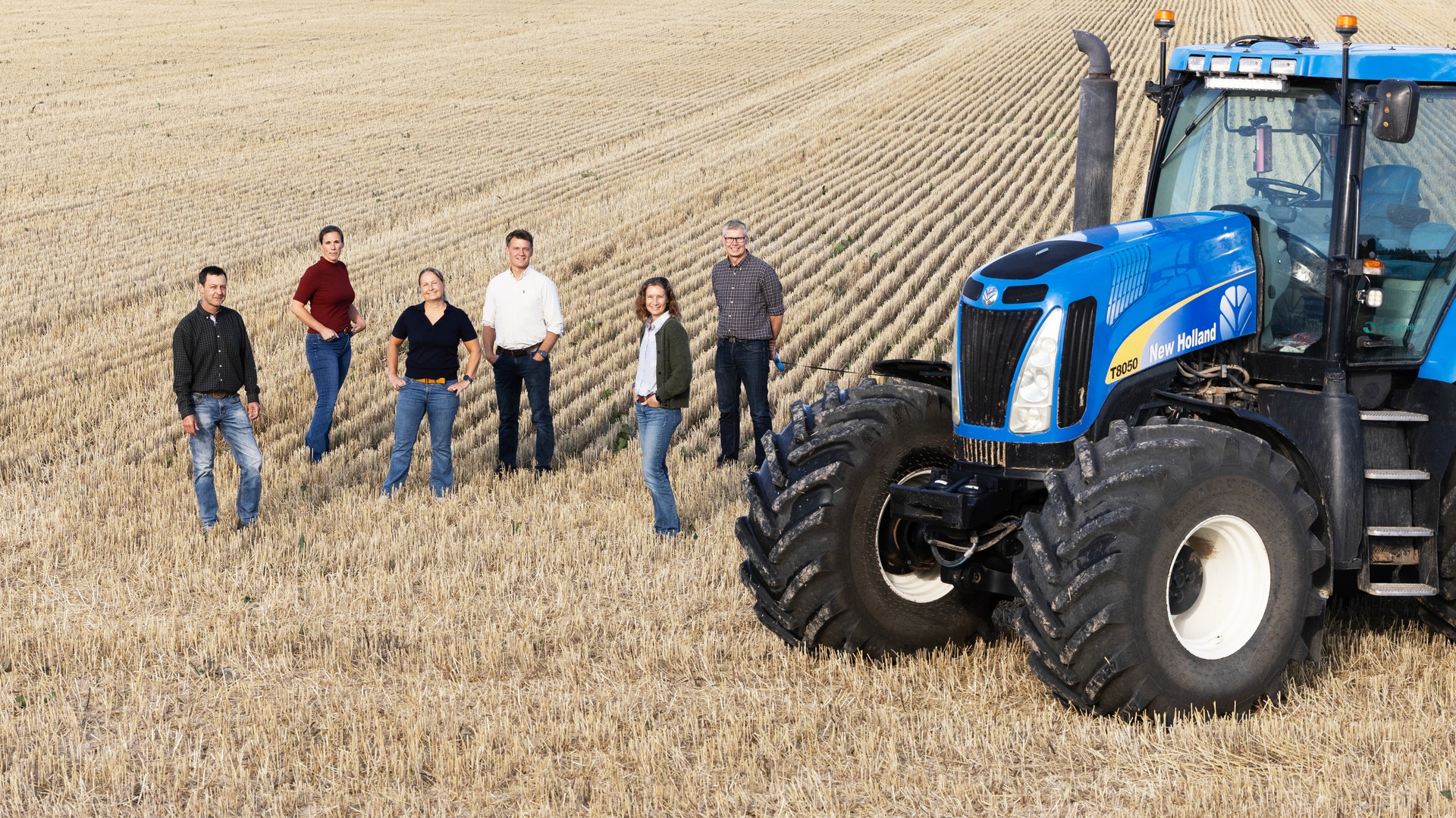 Sex personer står på ett skördat fält bredvid en stor blå traktor.