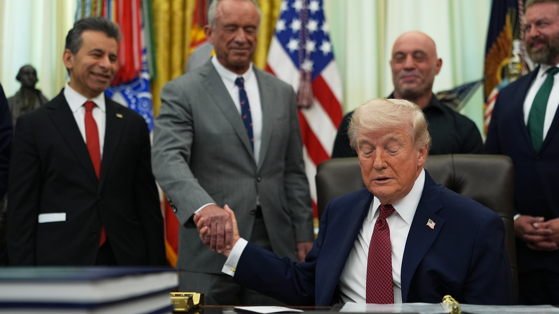 En man i mörkblå kostym och röd slips sitter vid ett skrivbord inomhus. Bakom honom står flera män i kostymer, och en amerikansk flagga syns i bakgrunden.