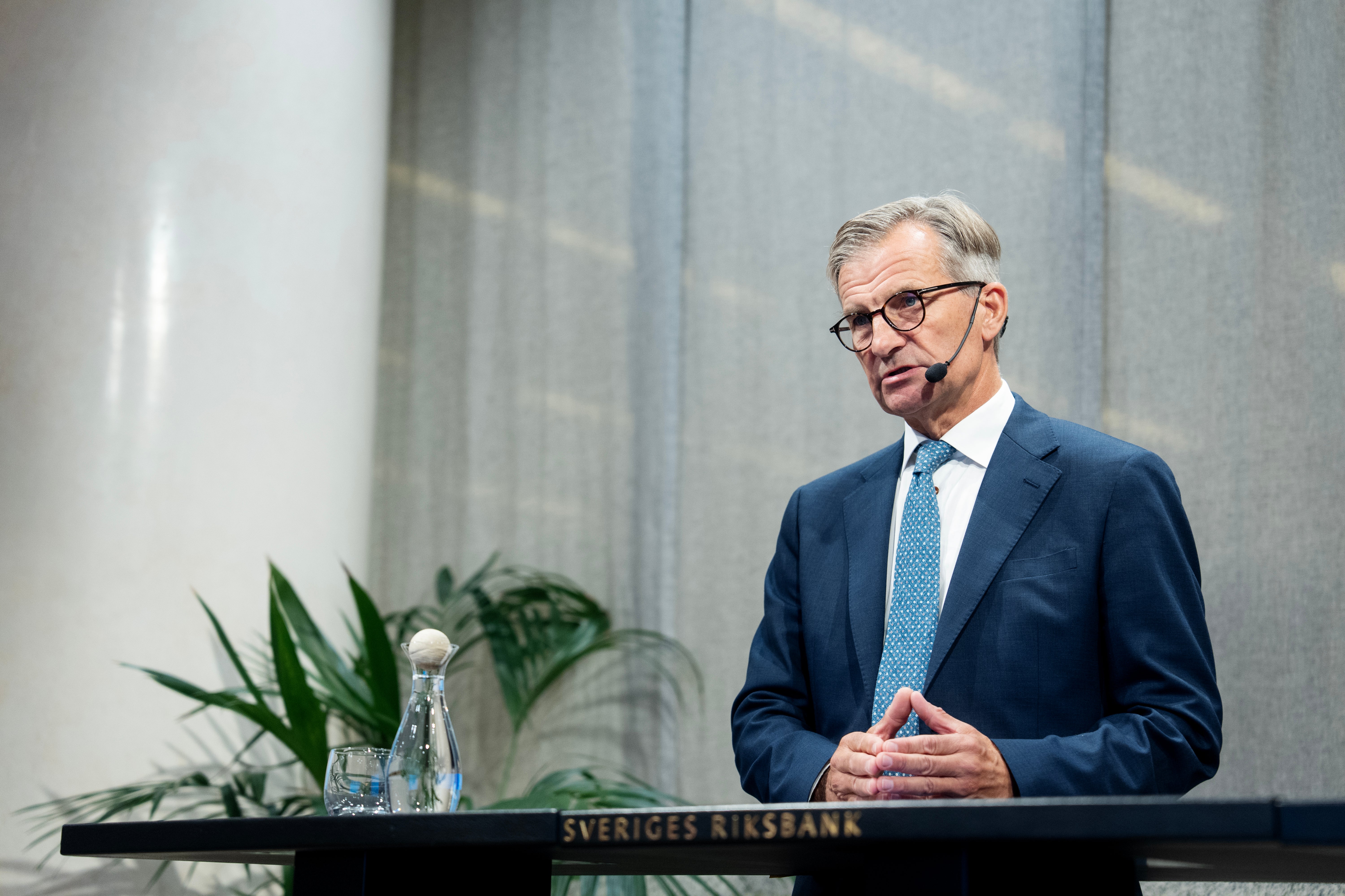En man i blå kostym och glasögon står vid ett podium märkt Sveriges Riksbank och gestikulerar medan han talar. En växt och en vattenkaraff syns på podiet bakom honom.
