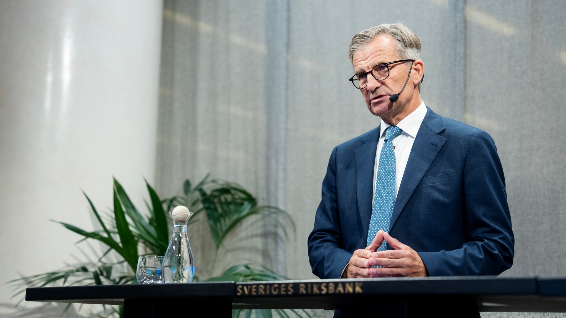 En man i blå kostym och glasögon står vid ett podium märkt Sveriges Riksbank och gestikulerar medan han talar. En växt och en vattenkaraff syns på podiet bakom honom.