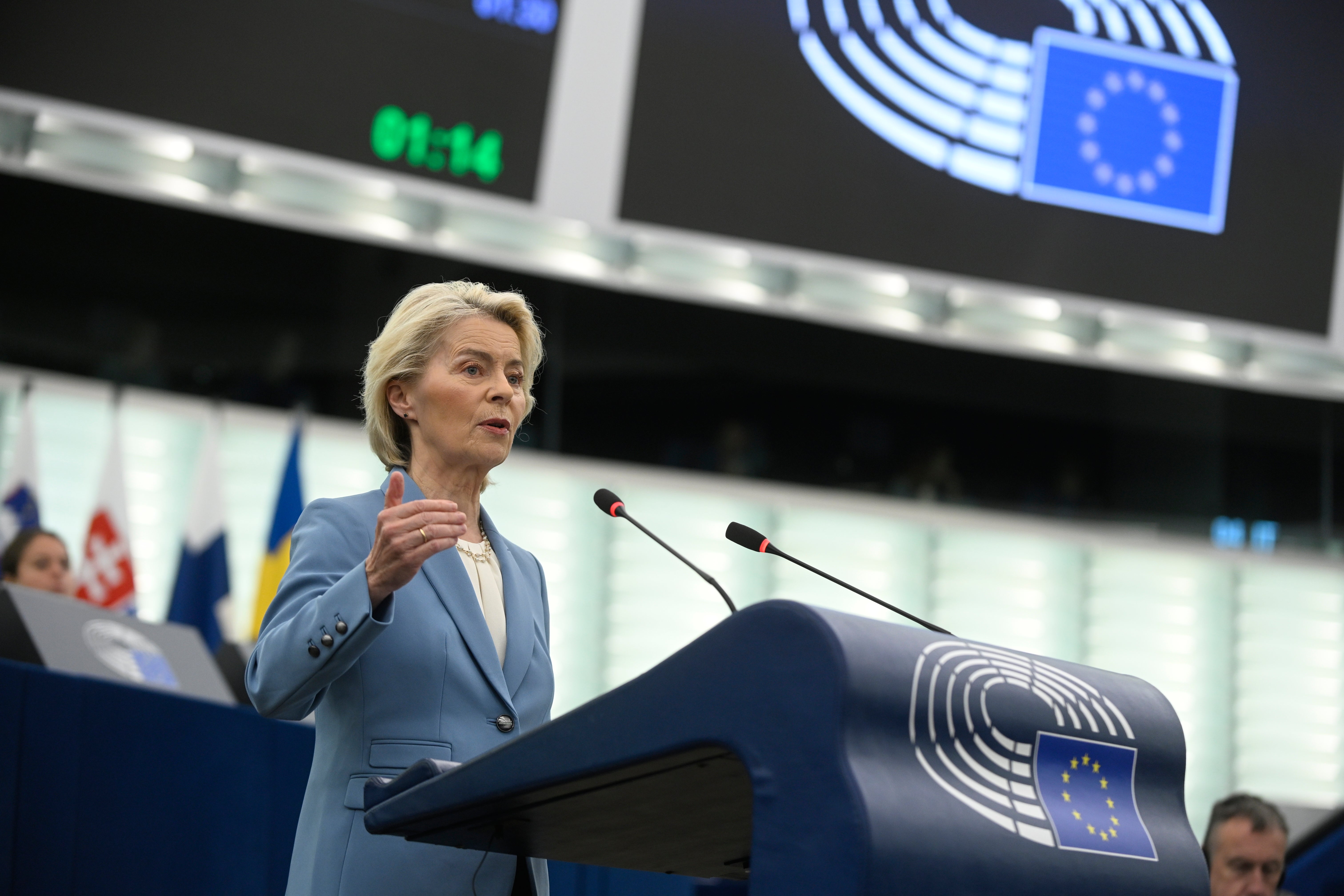 En kvinna i blå kavaj talar vid ett podium i en sal med EU-flaggor.