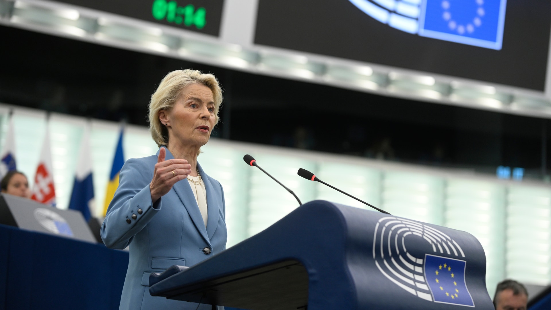 En kvinna i blå kavaj talar vid ett podium i en sal med EU-flaggor.
