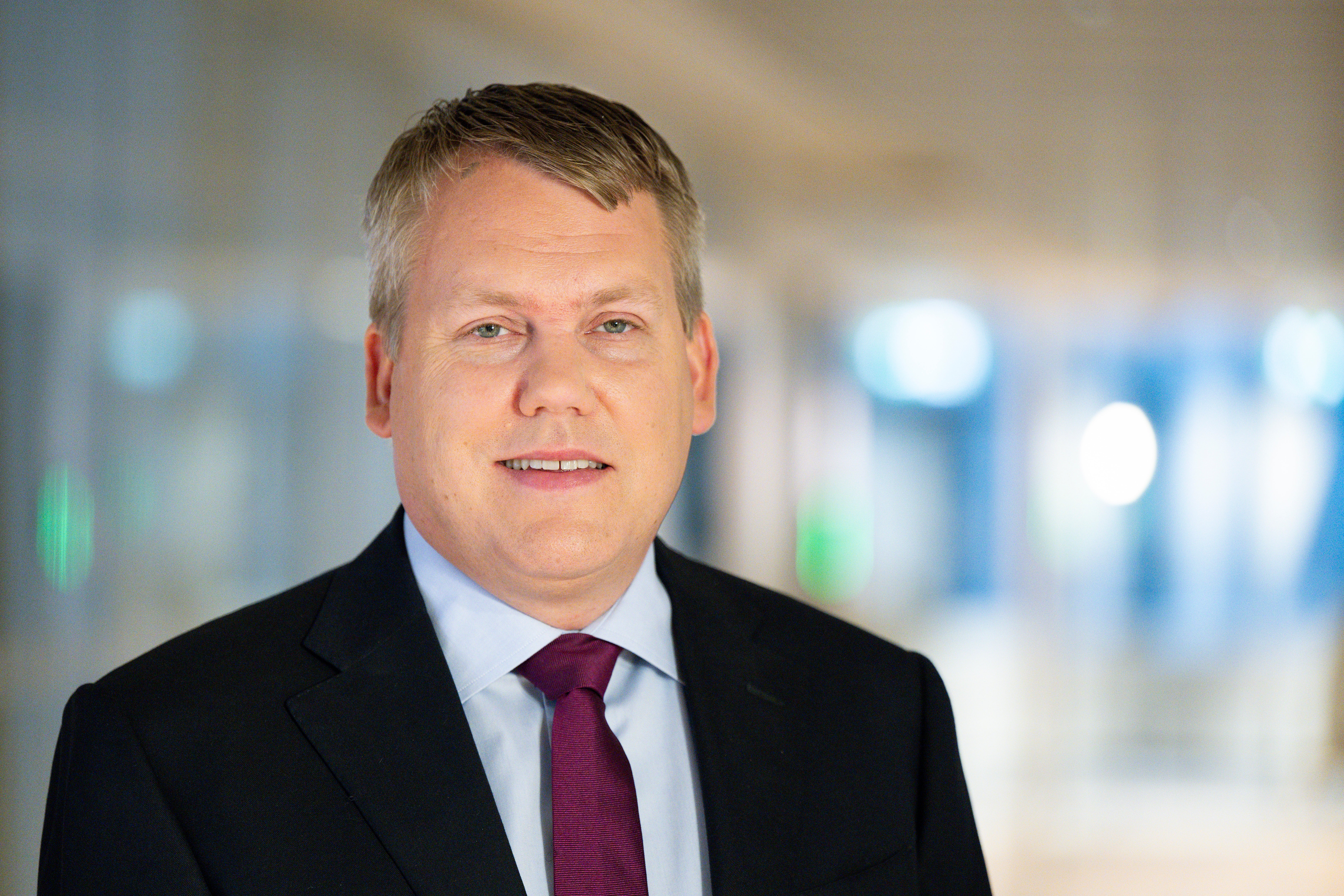 Porträttfoto av en man i svart kostym och röd slips mot en suddig kontorsbackgrund.