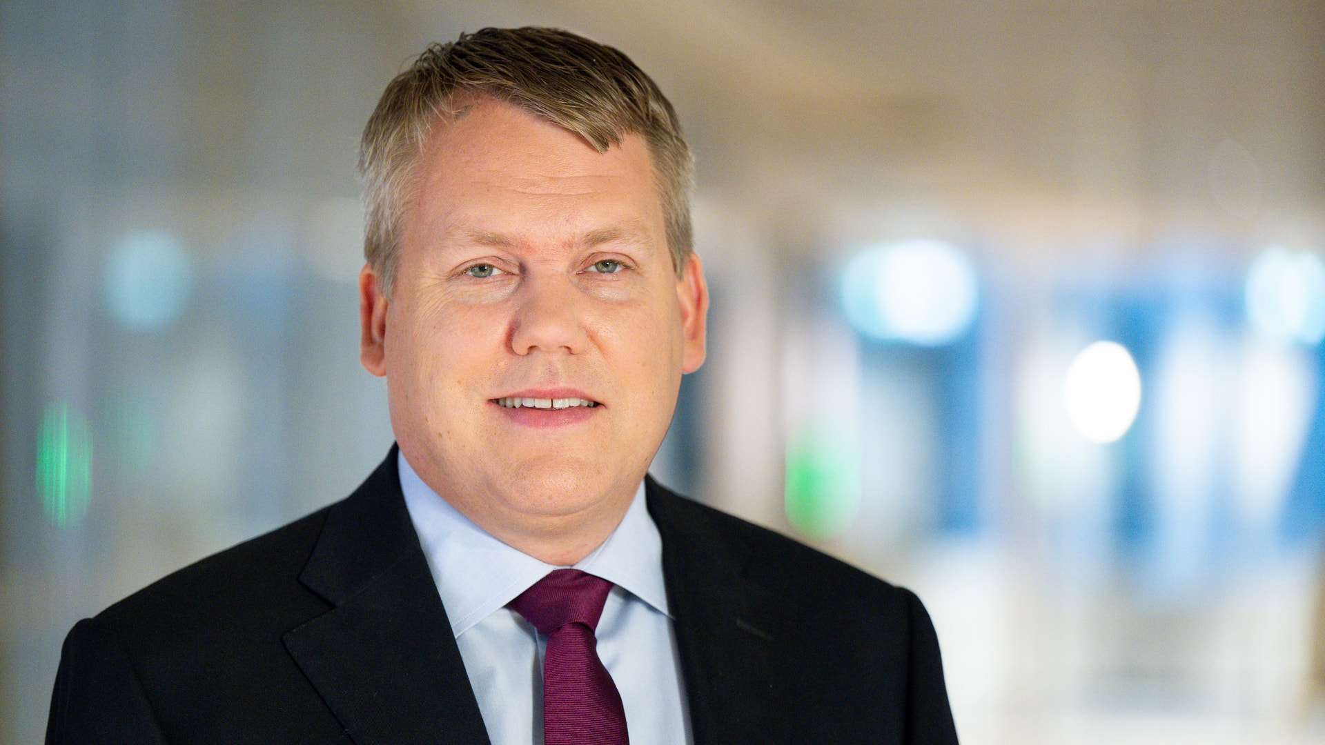 Porträttfoto av en man i svart kostym och röd slips mot en suddig kontorsbackgrund.