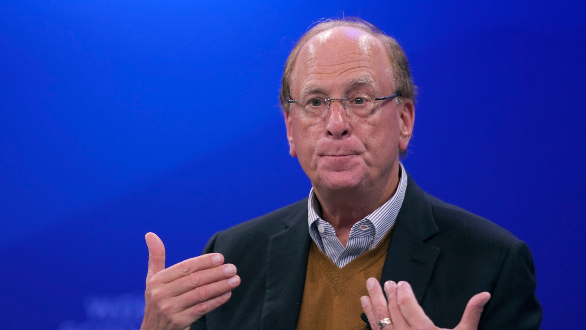 Larry Fink, en äldre man med glasögon och mörk kavaj, gestikulerar med båda händerna medan han pratar framför en blå bakgrund.