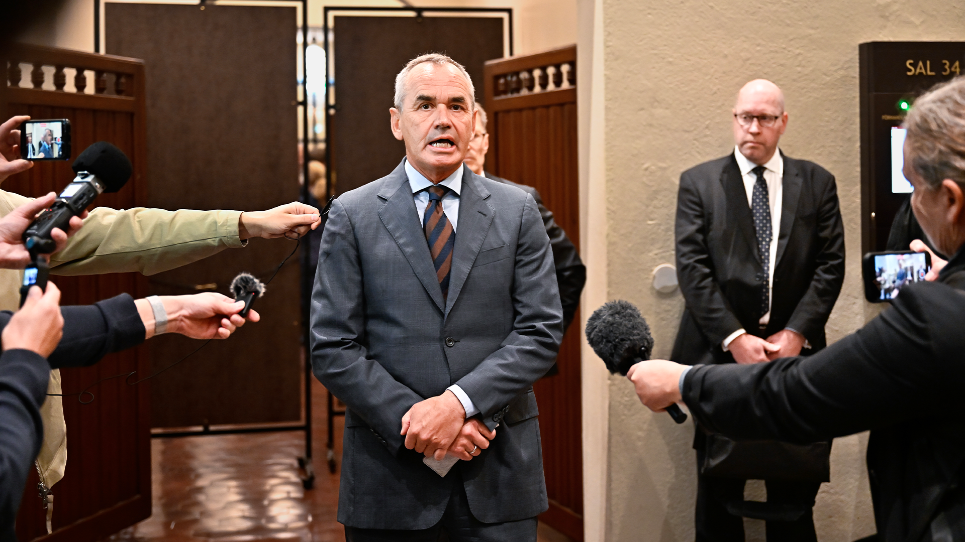 En man i blå kostym står omgiven av journalister med mikrofoner och kameror under en presskonferens.