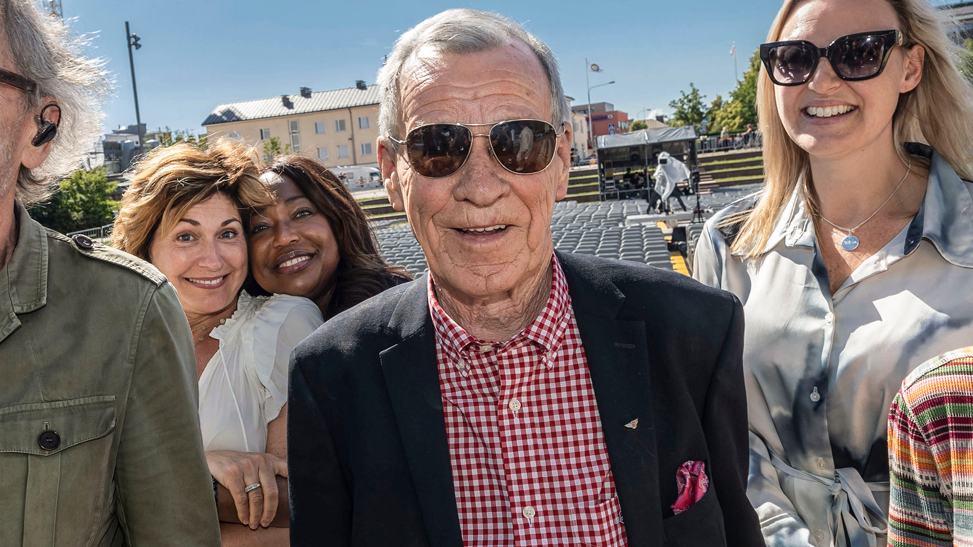 En grupp människor står tillsammans och ler mot kameran på en solig dag utomhus. En äldre man i mitten bär solglasögon och en marinblå blazer med röd-vit rutig skjorta.
