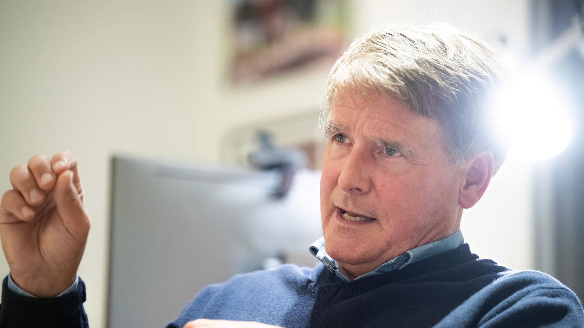 En äldre man med grått hår sitter och gestikulerar medan han talar. Han bär en blå och svart tröja.