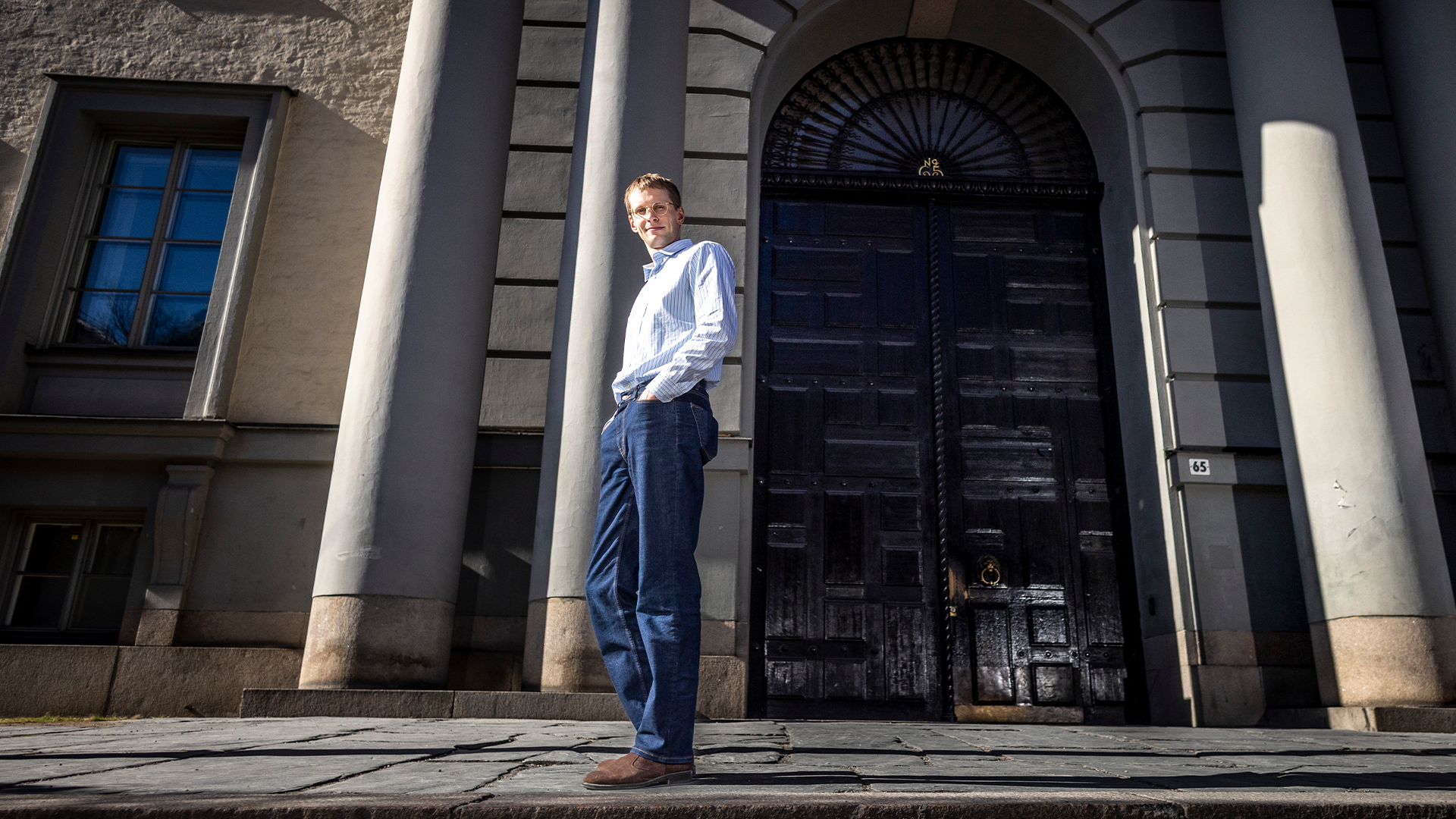 Man i vit skjorta och blå byxor står framför en klassisk byggnad med stora kolonner och mörk dörr.