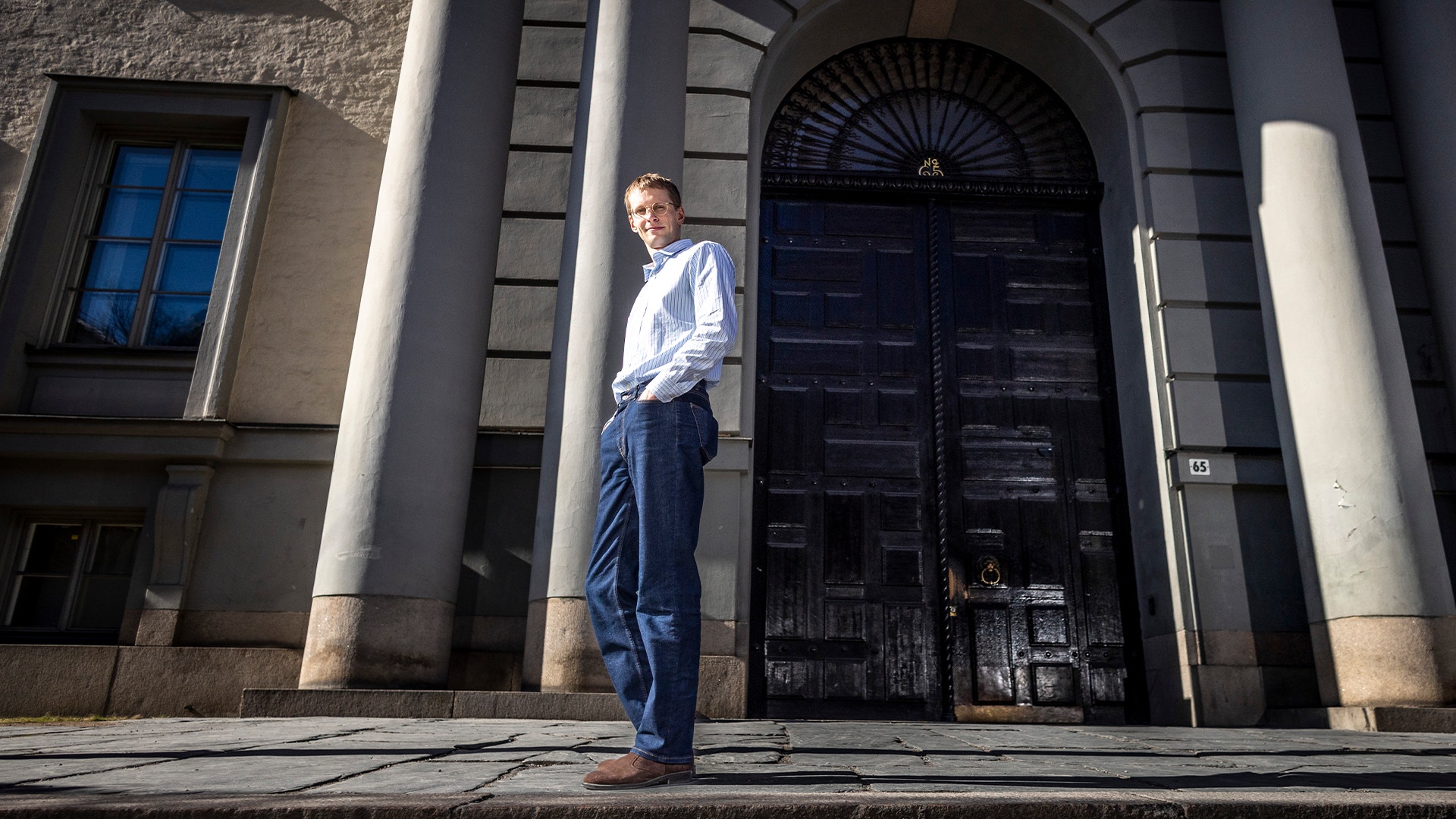Man i vit skjorta och blå byxor står framför en klassisk byggnad med stora kolonner och mörk dörr.
