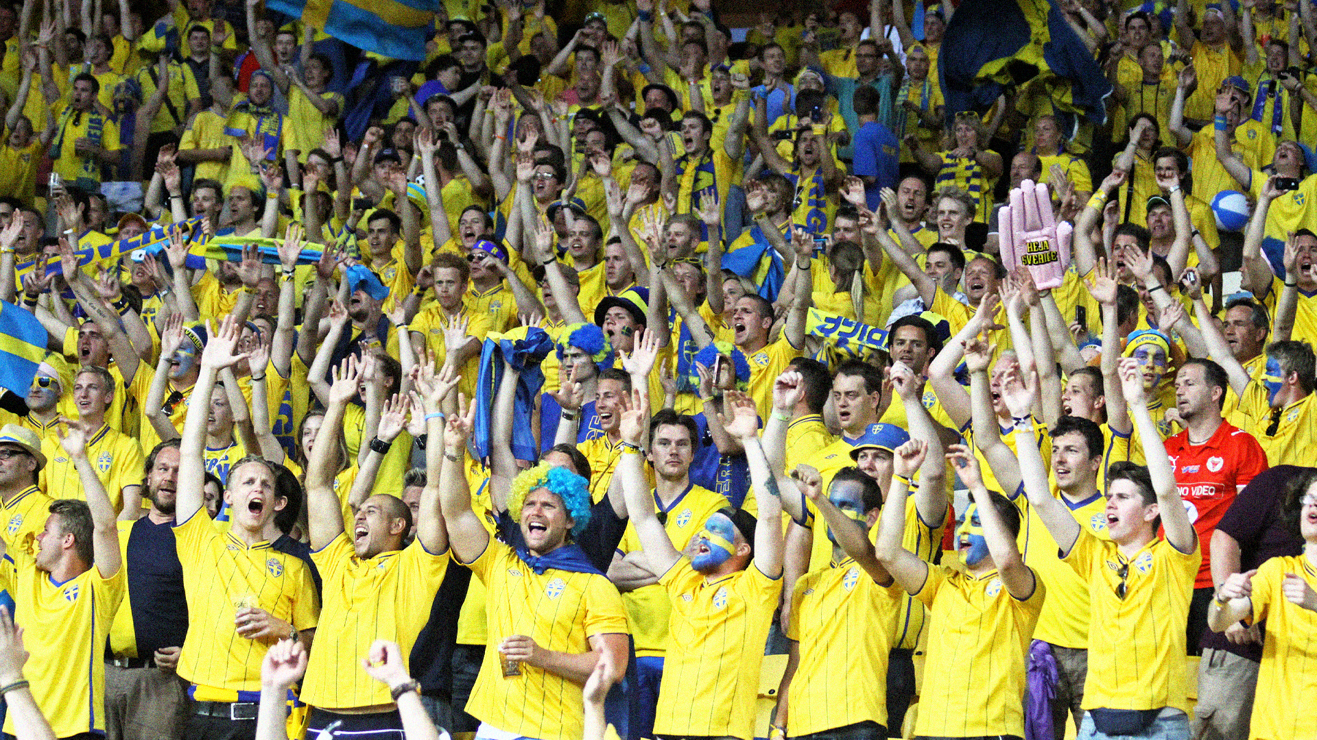 En stor grupp glada fotbollssupportrar i gula och blå kläder jublar och viftar på läktaren under en match.