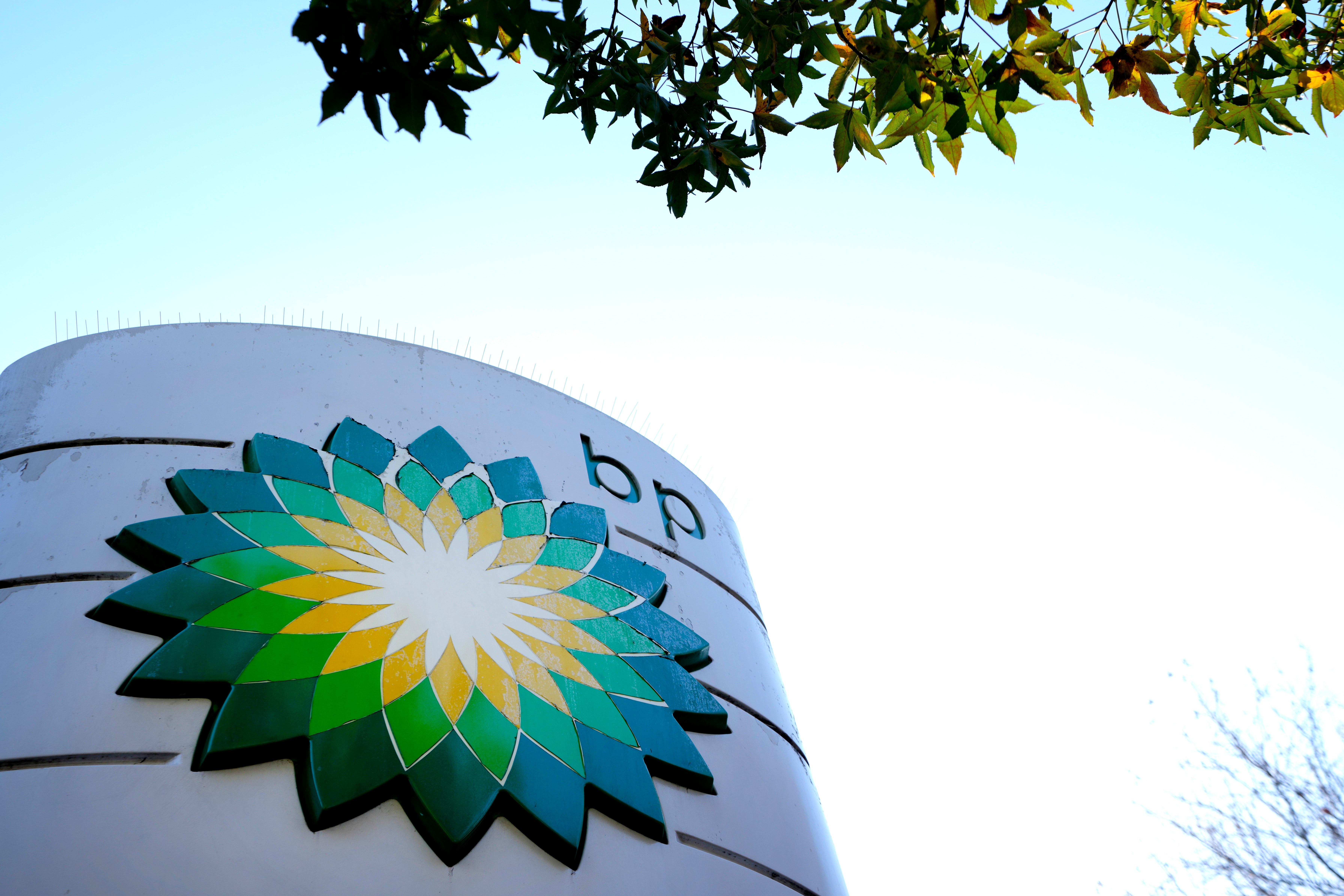 En stor cylindrisk behållare med BP:s blomsterlogo i grönt, gult och blått. Fotograferat underifrån mot blå himmel med träd synliga i bakgrunden.