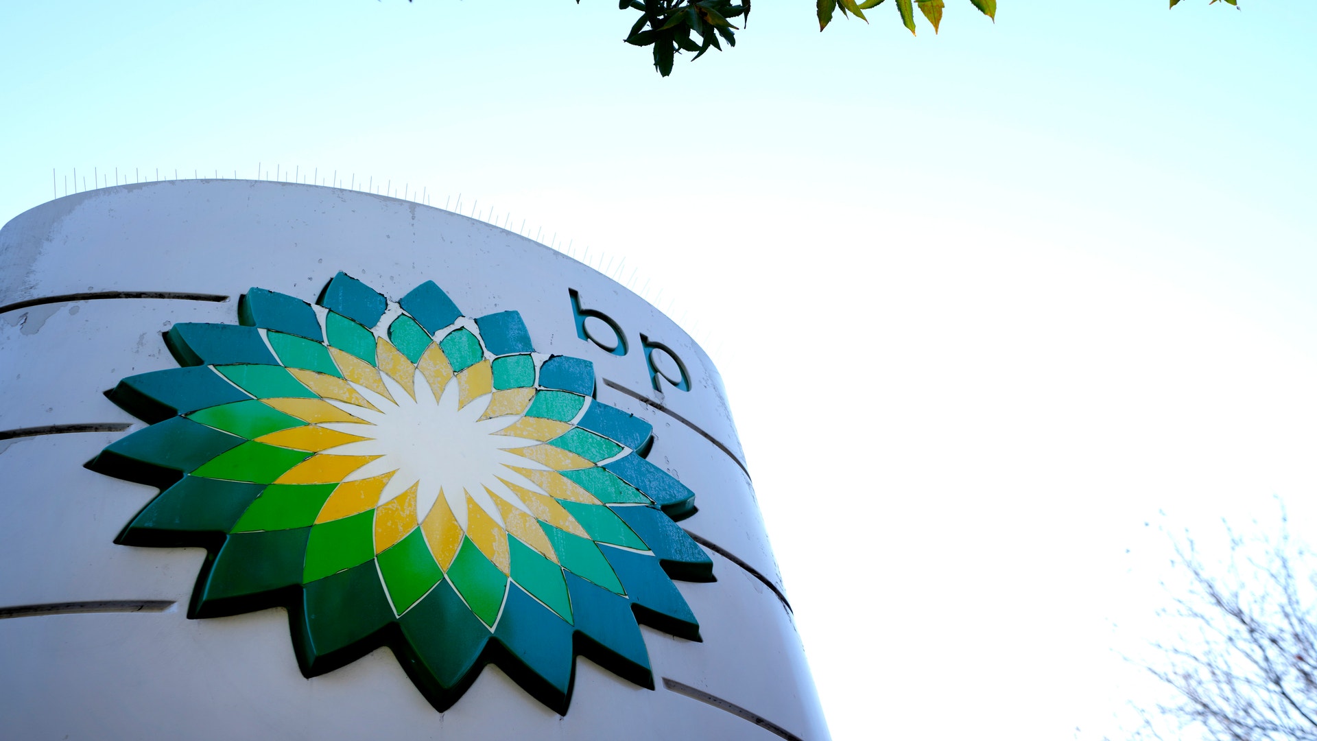 En stor cylindrisk behållare med BP:s blomsterlogo i grönt, gult och blått. Fotograferat underifrån mot blå himmel med träd synliga i bakgrunden.