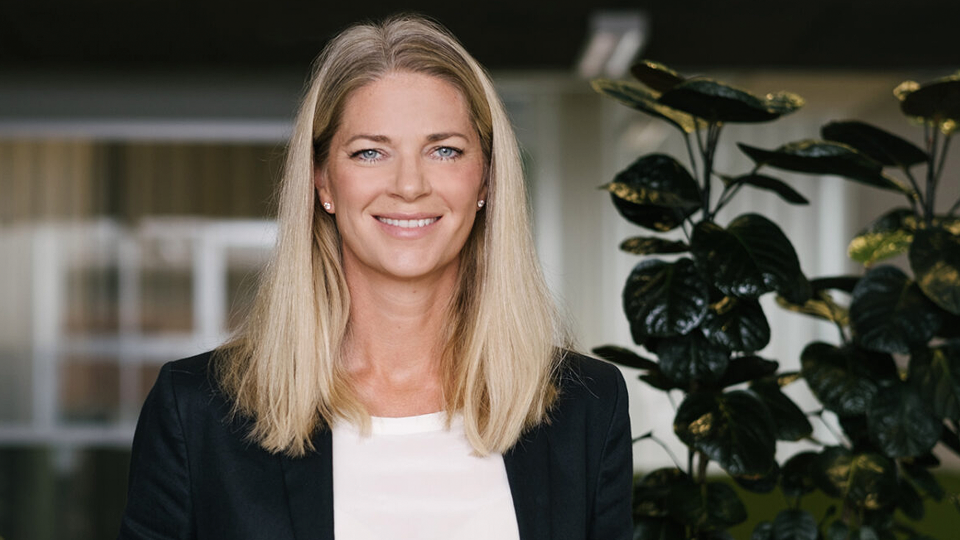 Porträtt av en kvinna med långt blont hår som bär en svart blazer och vit topp. Hon står framför en växt och ler mot kameran.