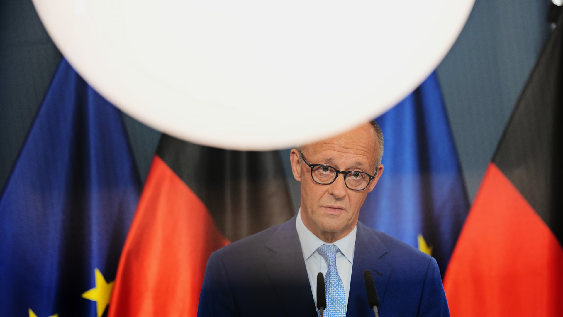 Man i kostym talar vid mikrofoner framför tyska och EU-flaggor.