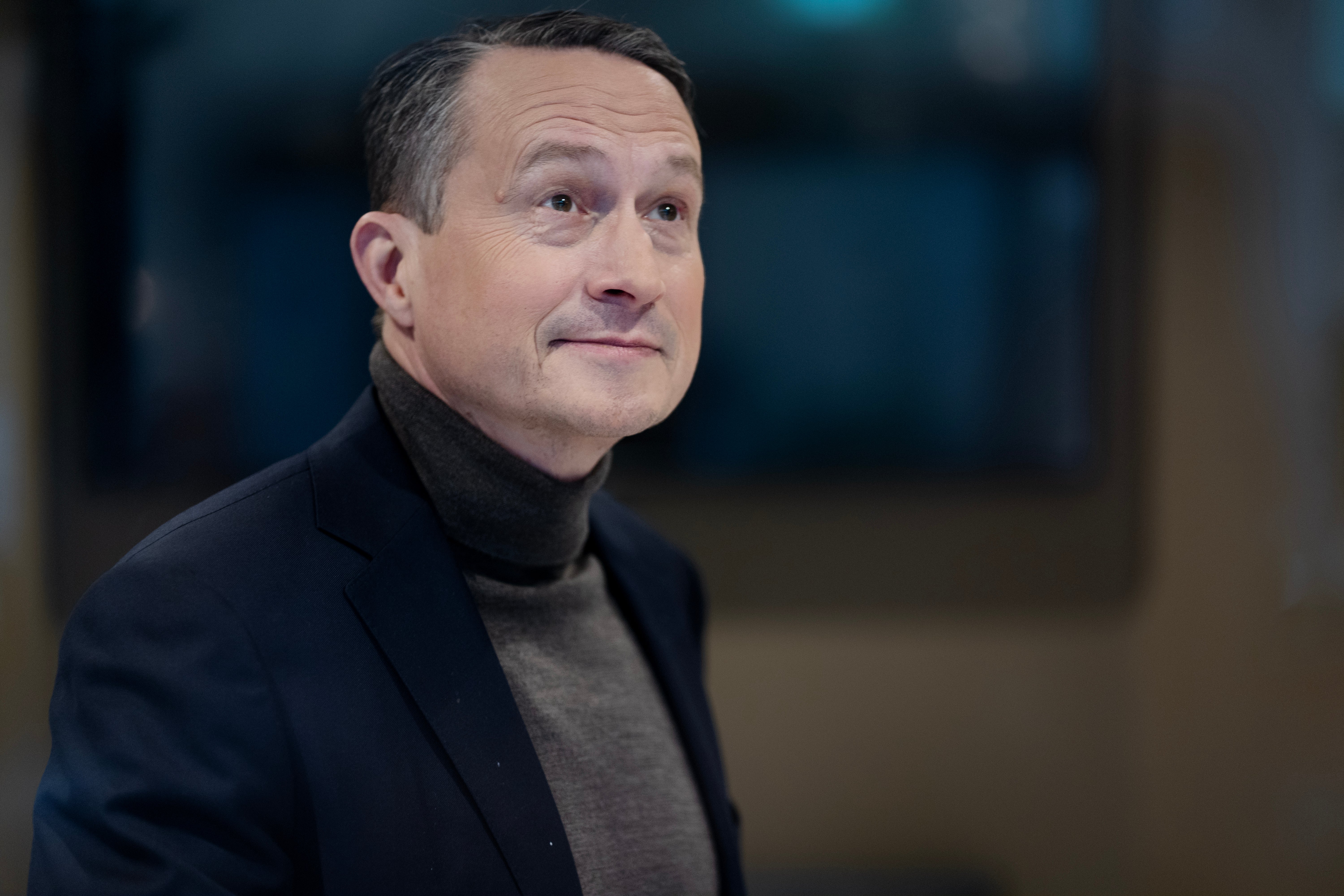 Porträtt av en man i mörk blazer och grå polotröja mot en suddig bakgrund