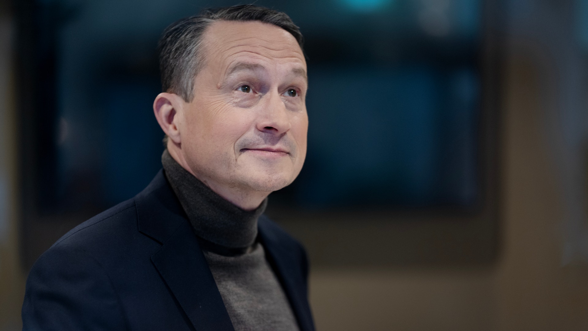 Porträtt av en man i mörk blazer och grå polotröja mot en suddig bakgrund