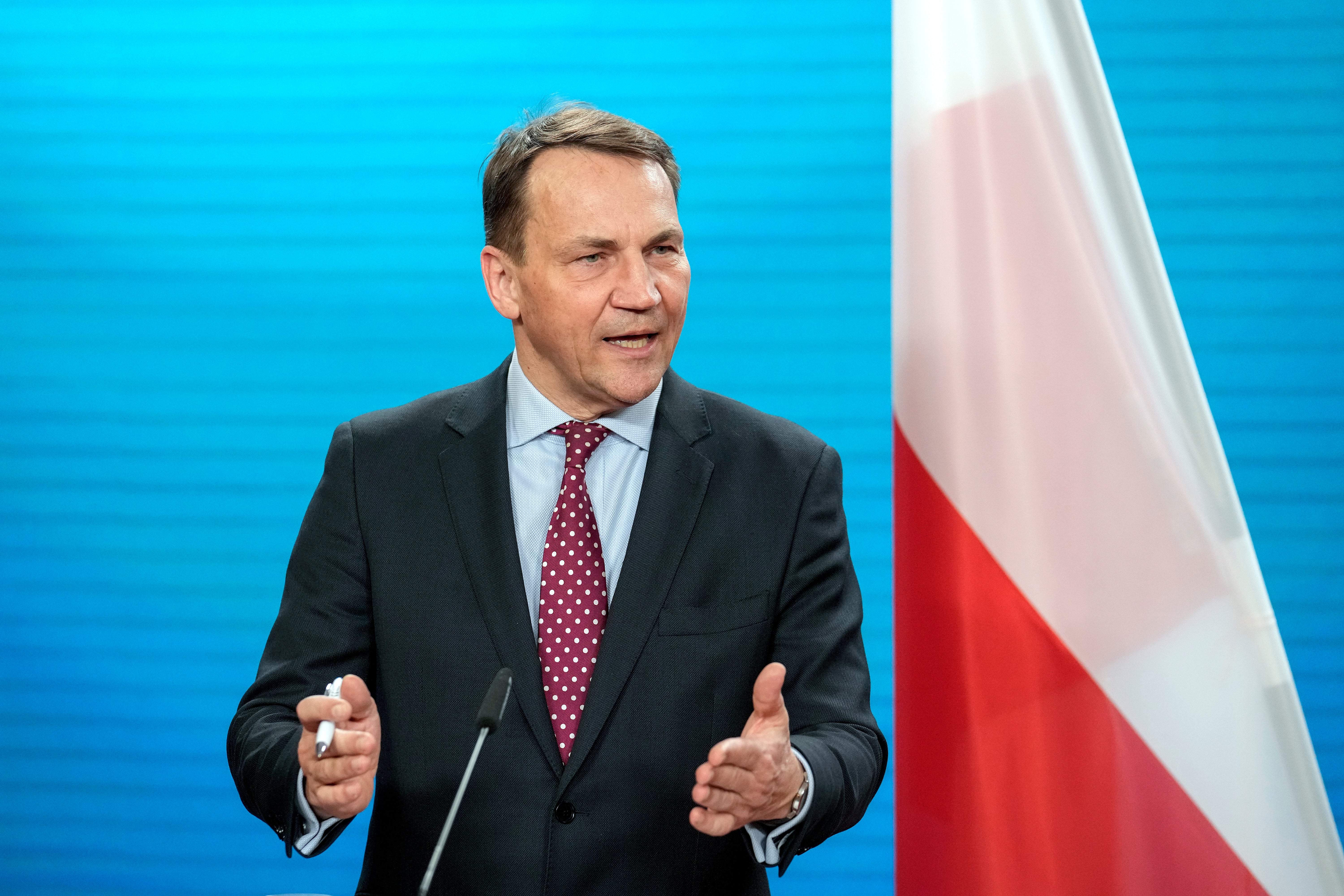 Polens utrikesminister Radosław Sikorski i mörk kostym talar och gestikulerar framför en polsk flagga.