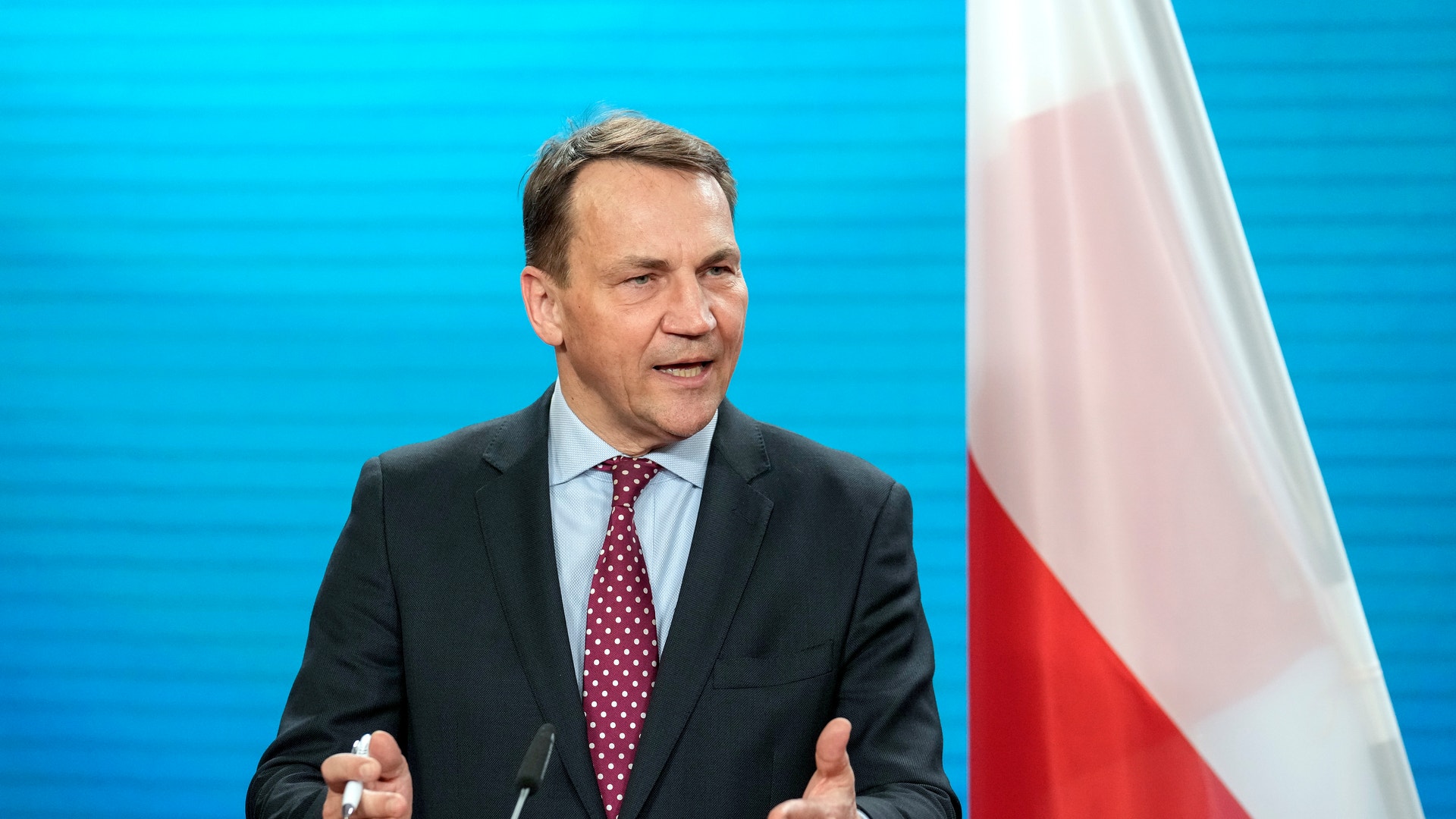 Polens utrikesminister Radosław Sikorski i mörk kostym talar och gestikulerar framför en polsk flagga.