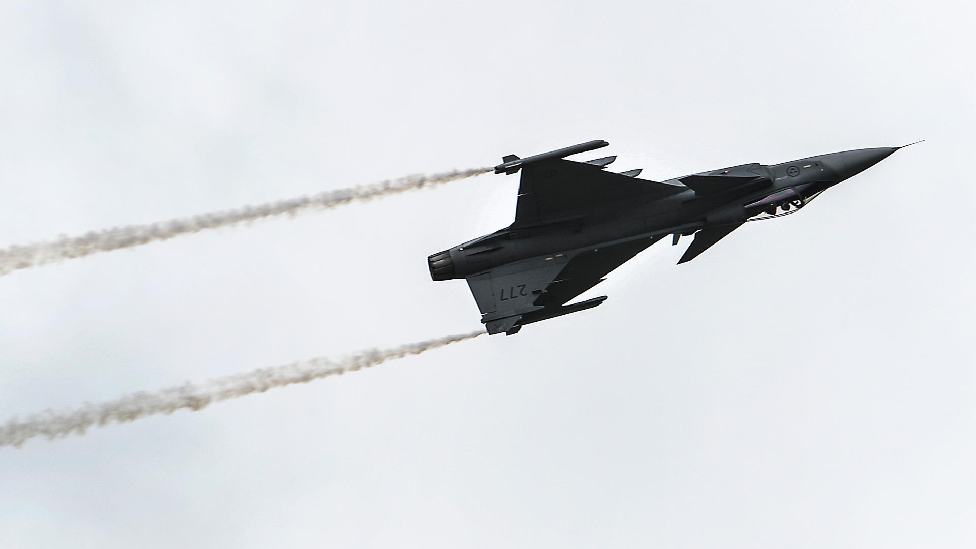 Ett svart stridsflygplan flyger mot en ljus himmel med två vita rökspår från motorerna bakom sig.