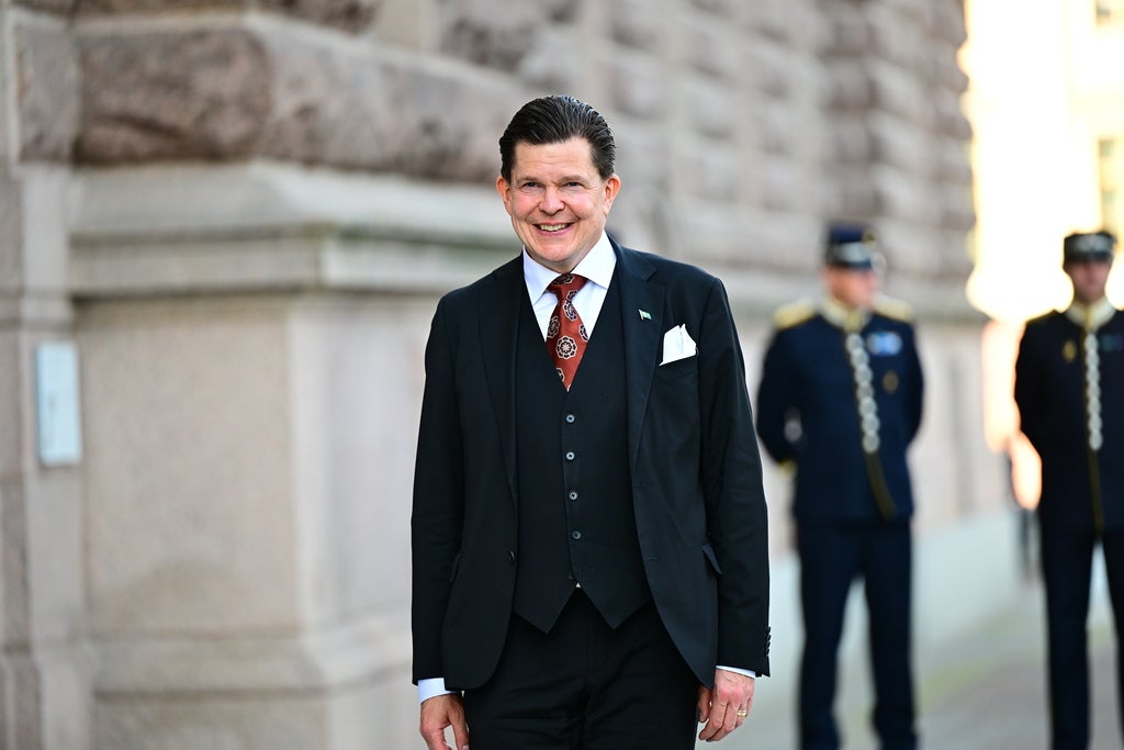 En leende man i kostym står utanför en stenbyggnad, med två uniformerade vakter i bakgrunden.