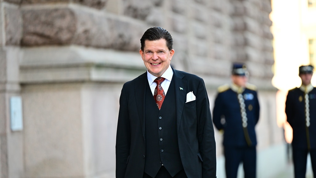 En leende man i kostym står utanför en stenbyggnad, med två uniformerade vakter i bakgrunden.