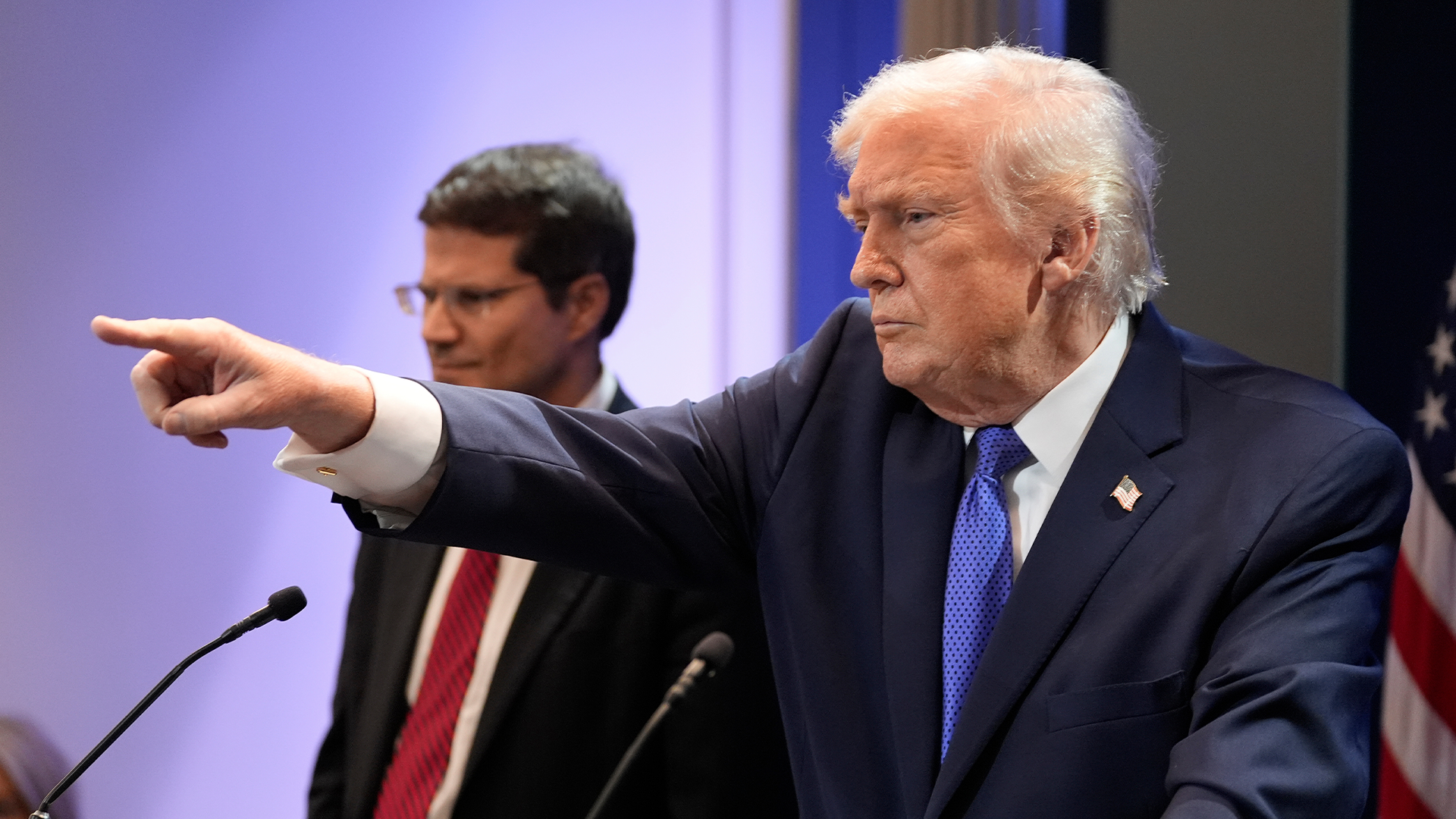 Äldre man i mörk kostym och blå slips pekar åt sidan under ett offentligt tal