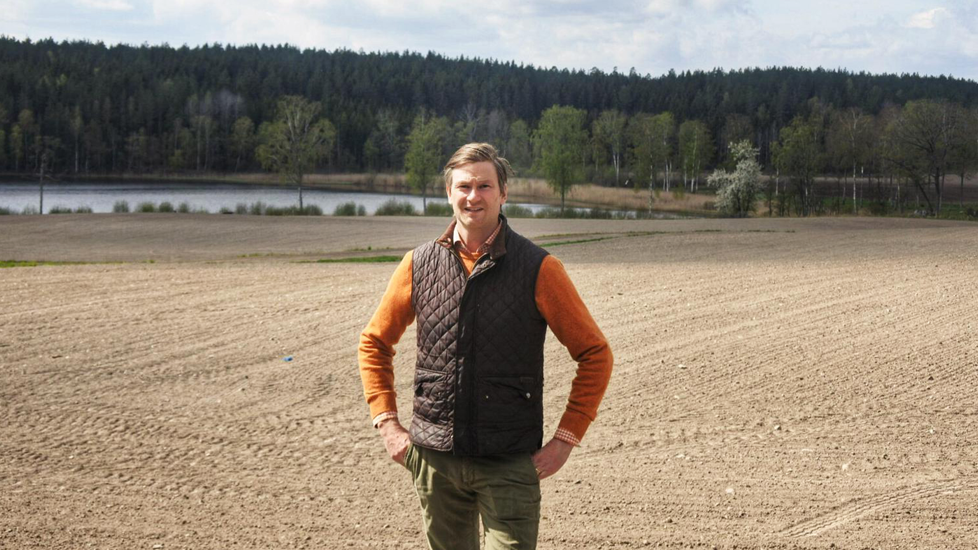 Porträtt av en man som står på ett plöjt åkerfält med skog och vatten i bakgrunden.
