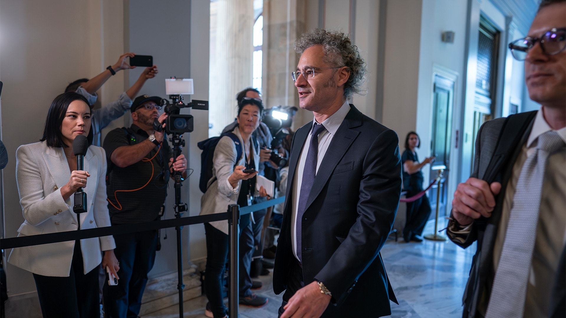 Alex Carp går genom entrén till Senaten i New York. Han omringas av reportrar.