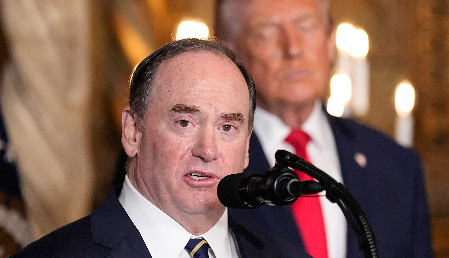 En man talar vid en mikrofon i en formell inomhusmiljö.