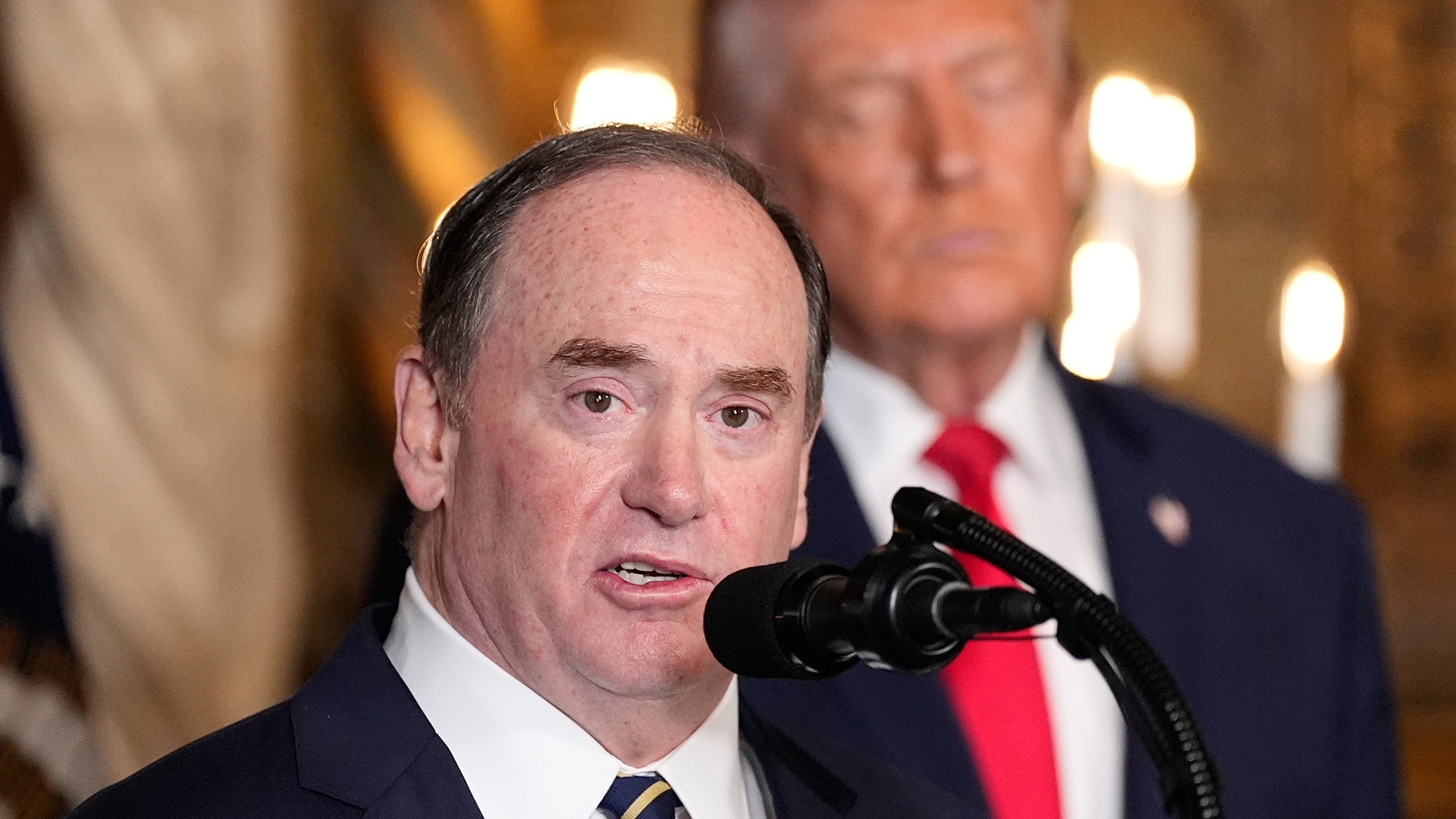 En man talar vid en mikrofon i en formell inomhusmiljö.