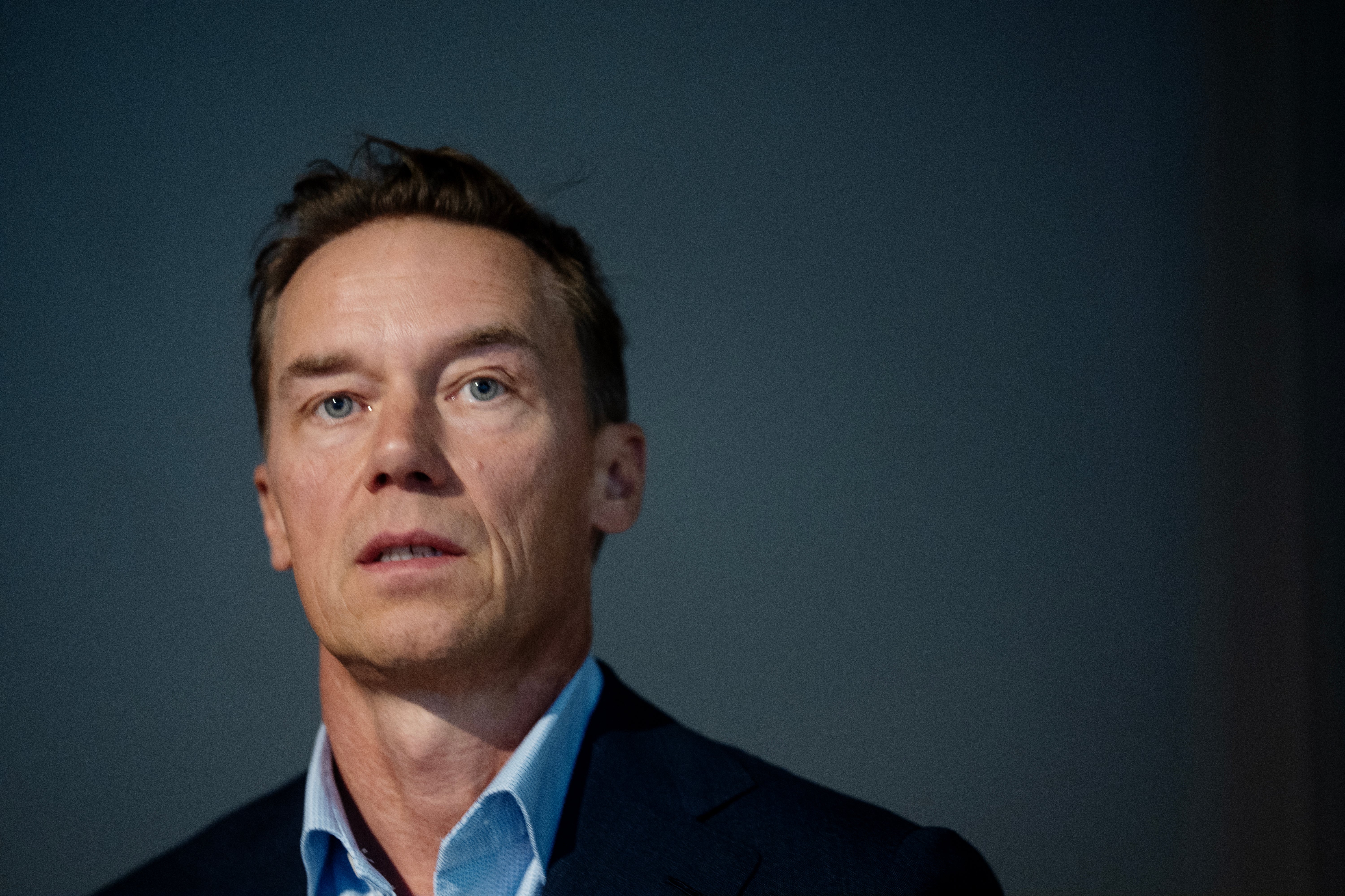 Porträtt av en man i blå skjorta och mörk kavaj mot blå bakgrund.