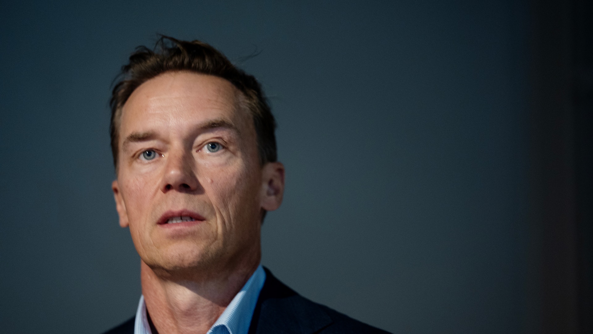 Porträtt av en man i blå skjorta och mörk kavaj mot blå bakgrund.