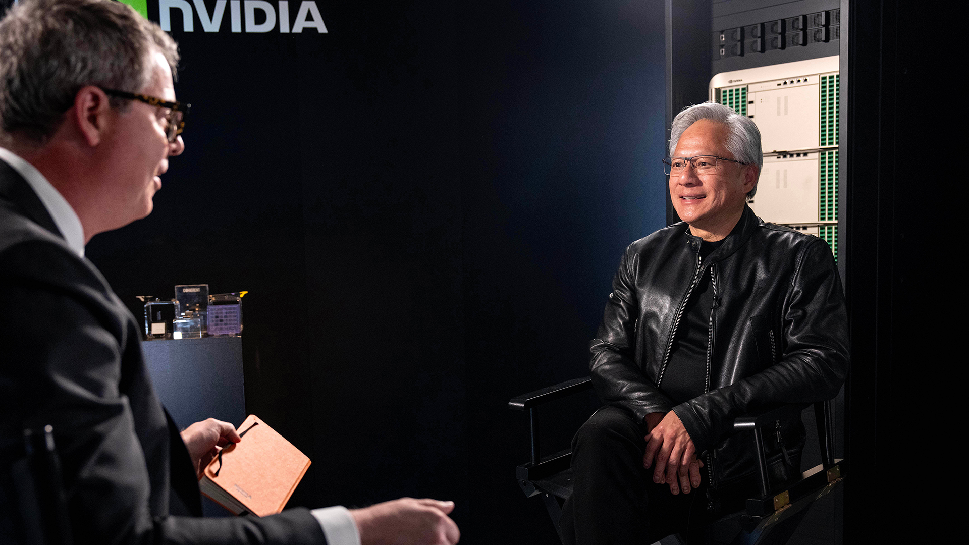 Två män i en intervjusituation. Mannen till vänster bär glasögon och mörkdräkt, mannen till höger bär svart läderrock. I bakgrunden syns Nvidia-logotypen och datorhårdvara.