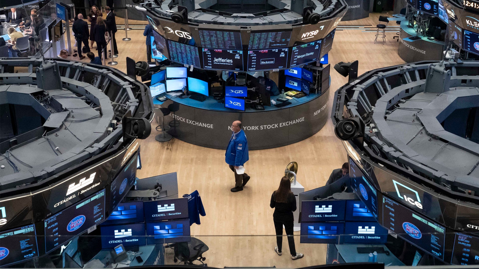 Handelsgolvet på New York Stock Exchange sett från ovan. Flera handlare i mörkblå och svarta kläder arbetar vid datorskärmar och handelsstationer. Stora blå informationsskärmar visar aktiedata runt omkring.