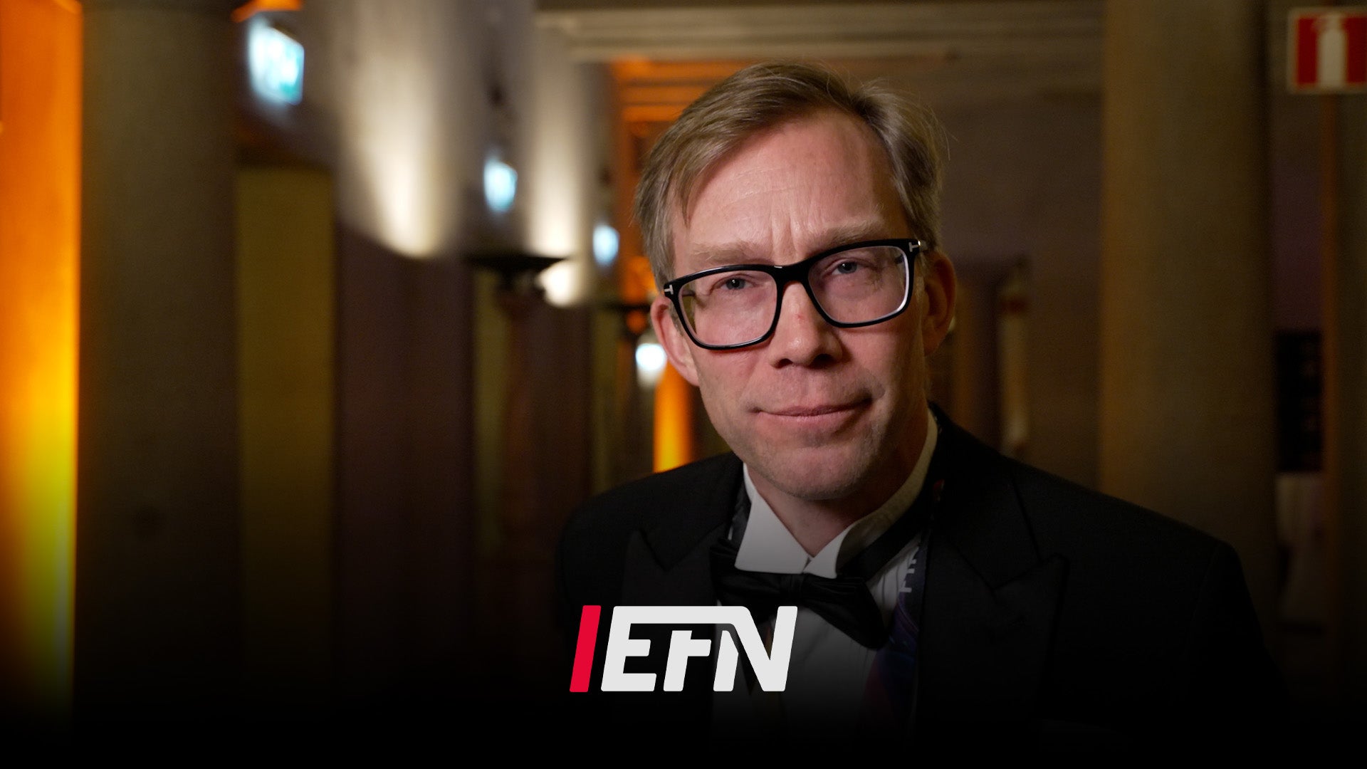 Man med glasögon håller en mikrofon inomhus i en korridor