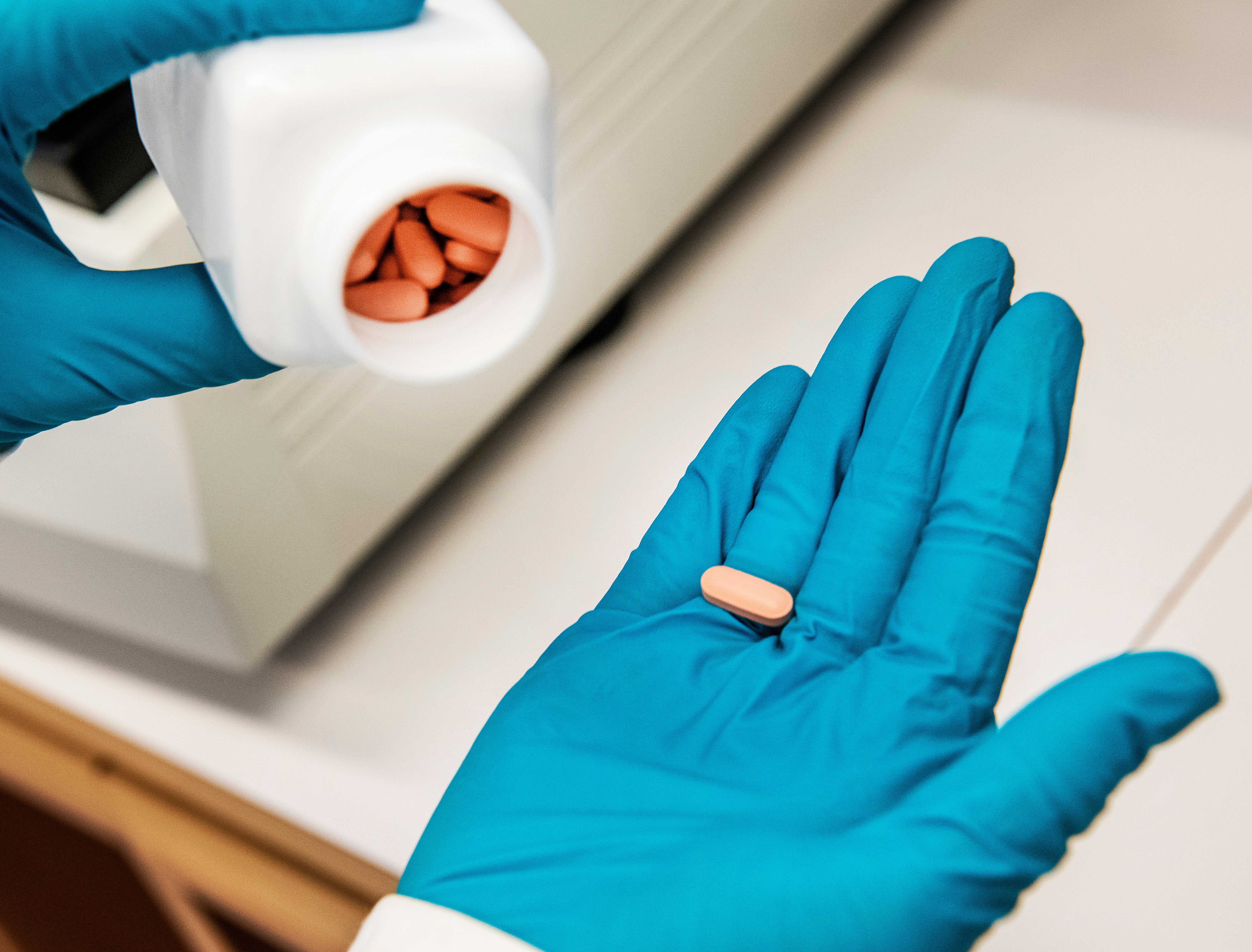 Person med blå medicinska handskar håller en orange tablett i handen. En vit behållare med fler tabletter syns i bakgrunden.