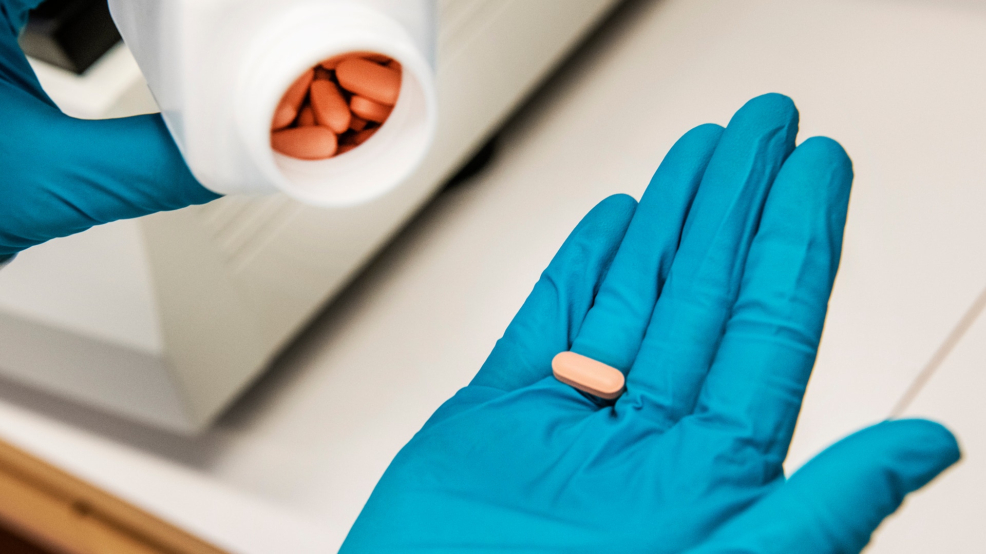Person med blå medicinska handskar håller en orange tablett i handen. En vit behållare med fler tabletter syns i bakgrunden.