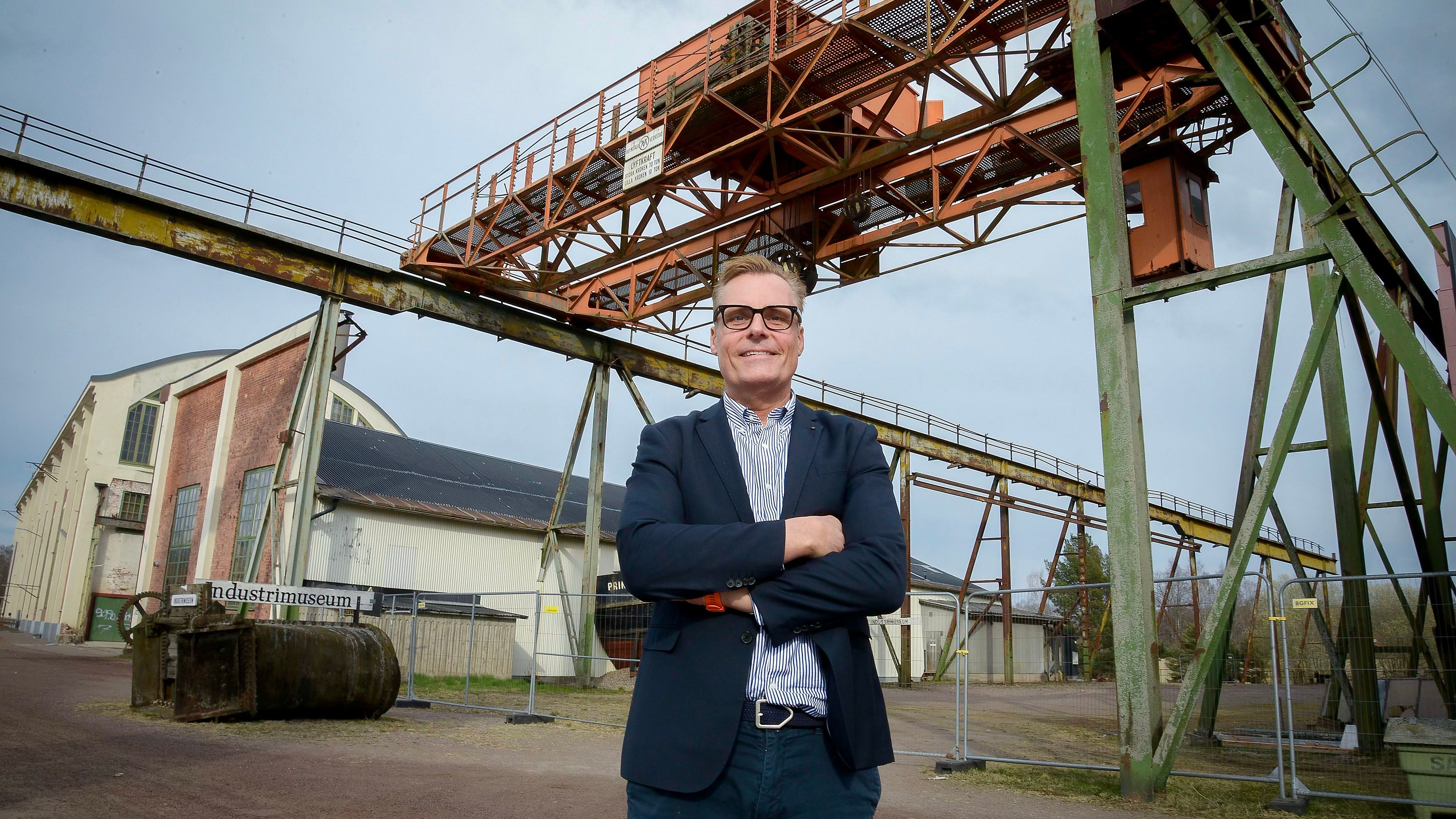 Man i mörk kostym och glasögon står framför en stor industrianläggning med kranstrukturer och metallkonstruktioner.