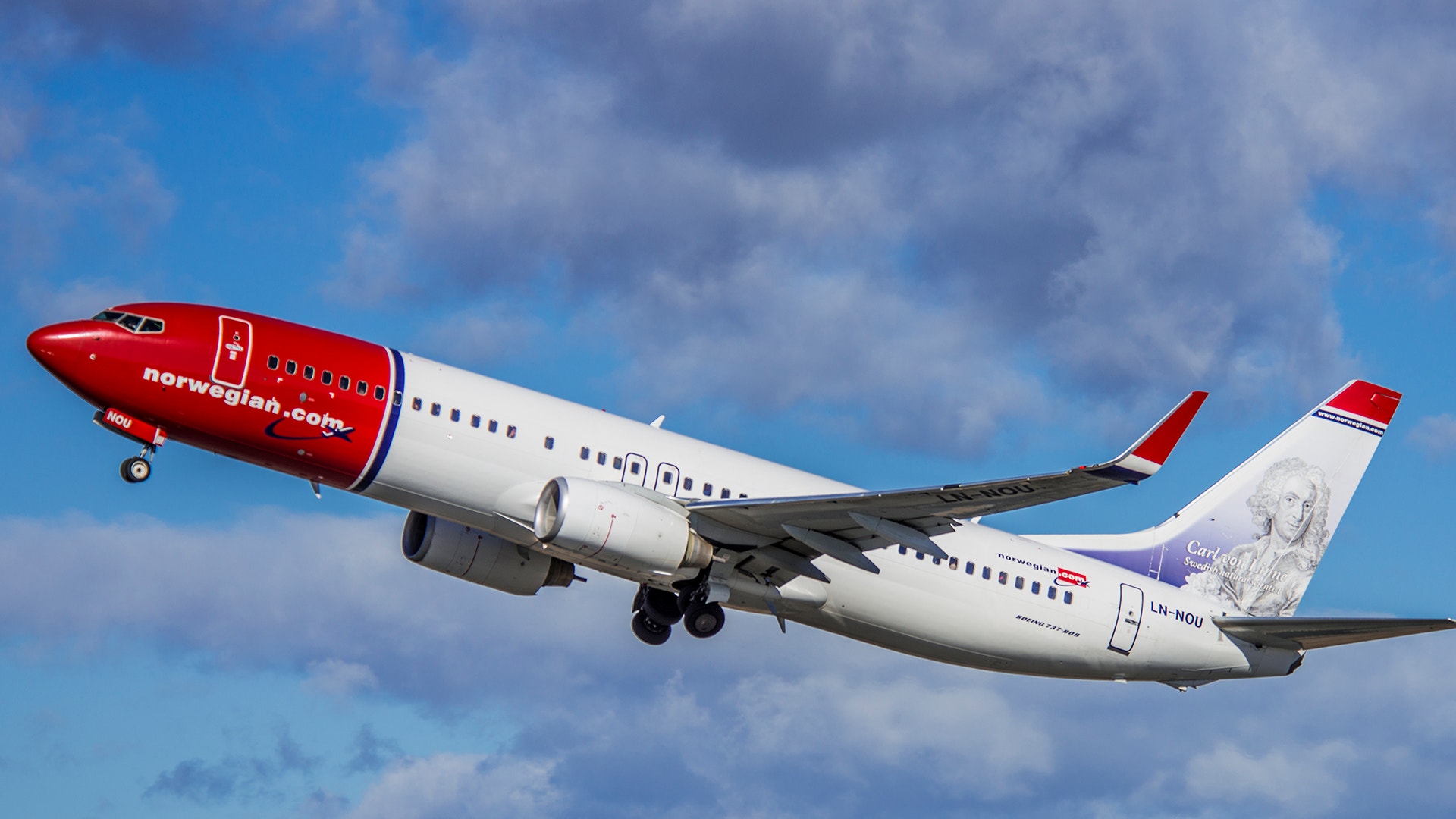 Ett rött och vitt Norwegian-flygplan i luften mot en blå himmel med vita moln.