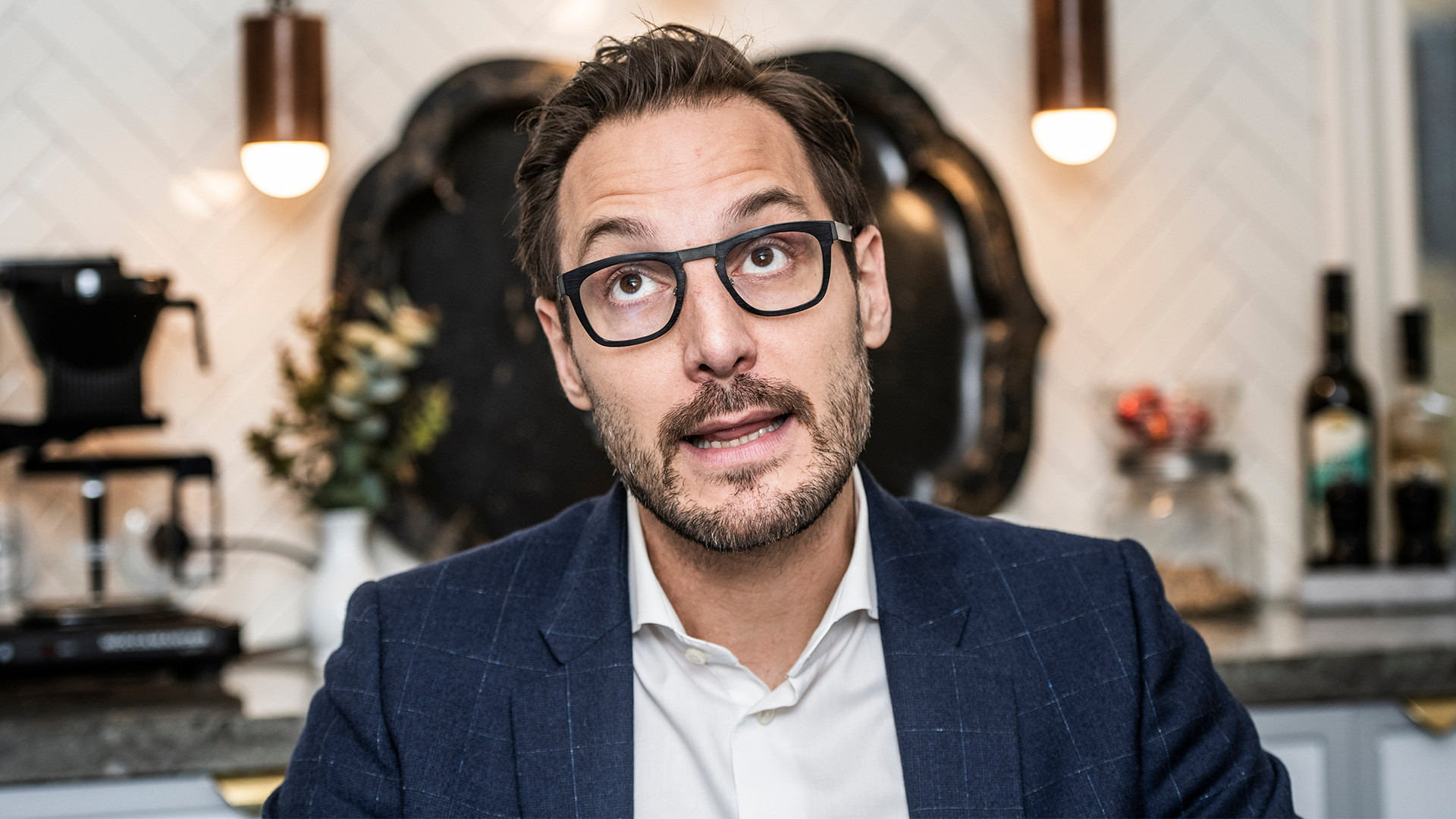 Porträtt av en man med glasögon i blå kavaj och vit skjorta, fotograferad i en modern inomhusmiljö.