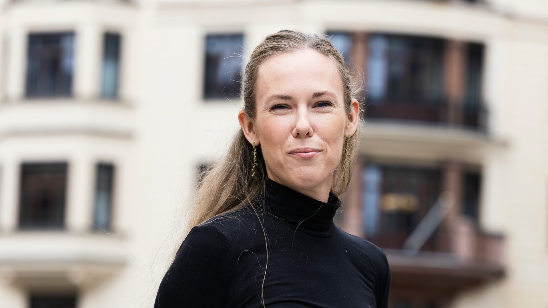 Porträtt av en kvinna med långt ljust hår, iklädd svart polotröja, fotograferad framför en beige byggnad med fönster.