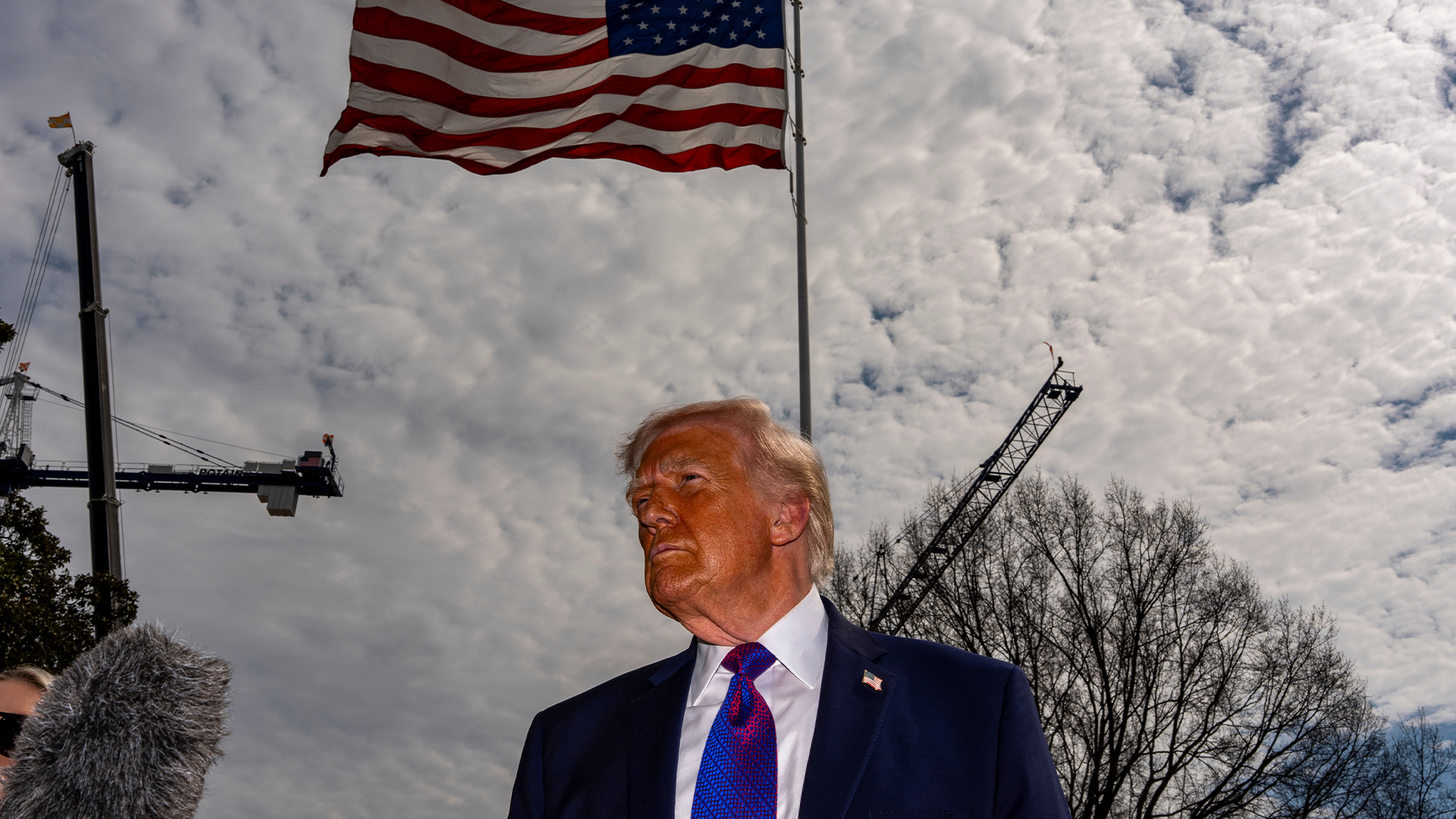 Man i mörk kostym och blå slips framför amerikansk flagga och molnig himmel.