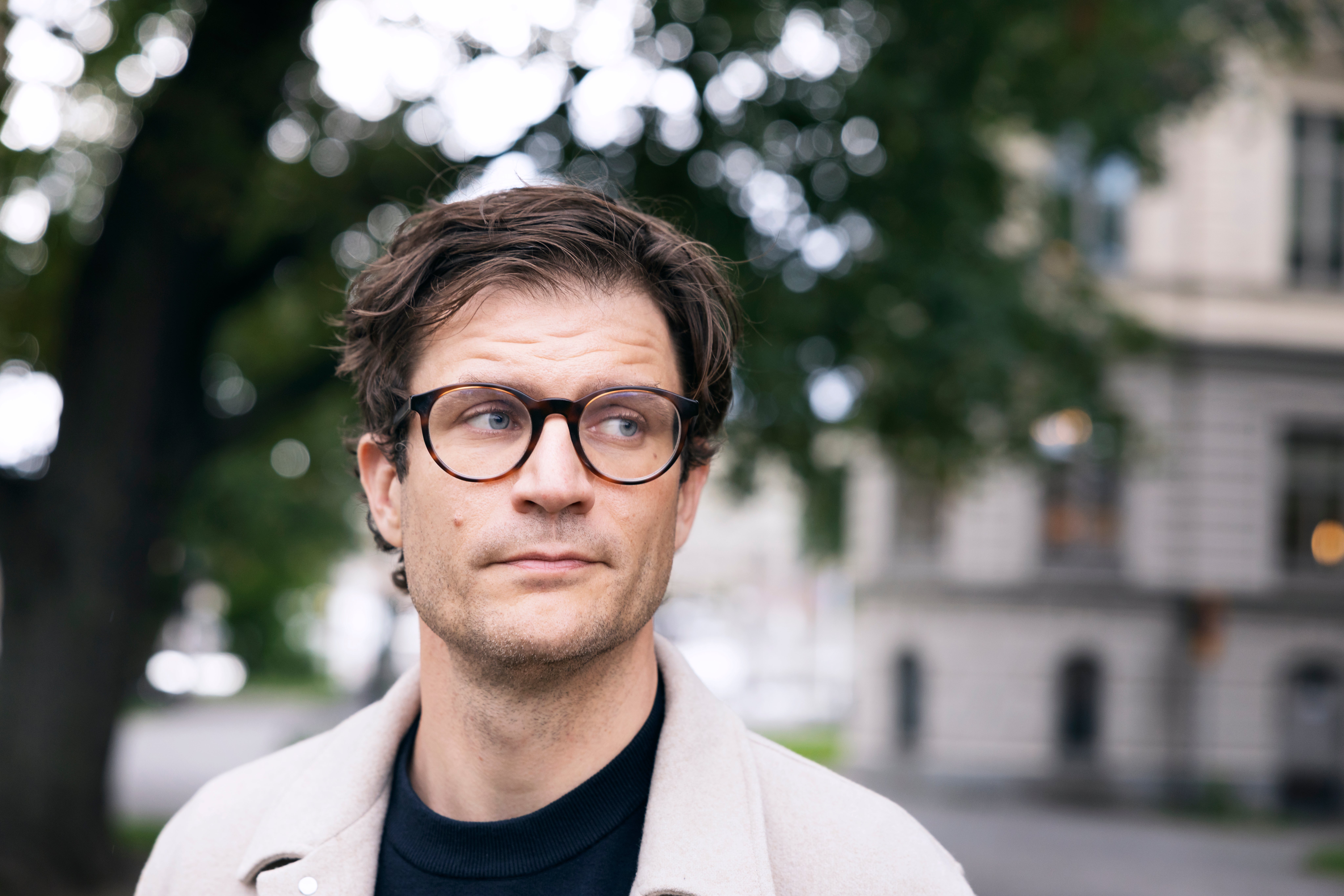 Porträtt av en man med glasögon och ljus jacka som står utomhus framför ett byggnadsliknande objekt.