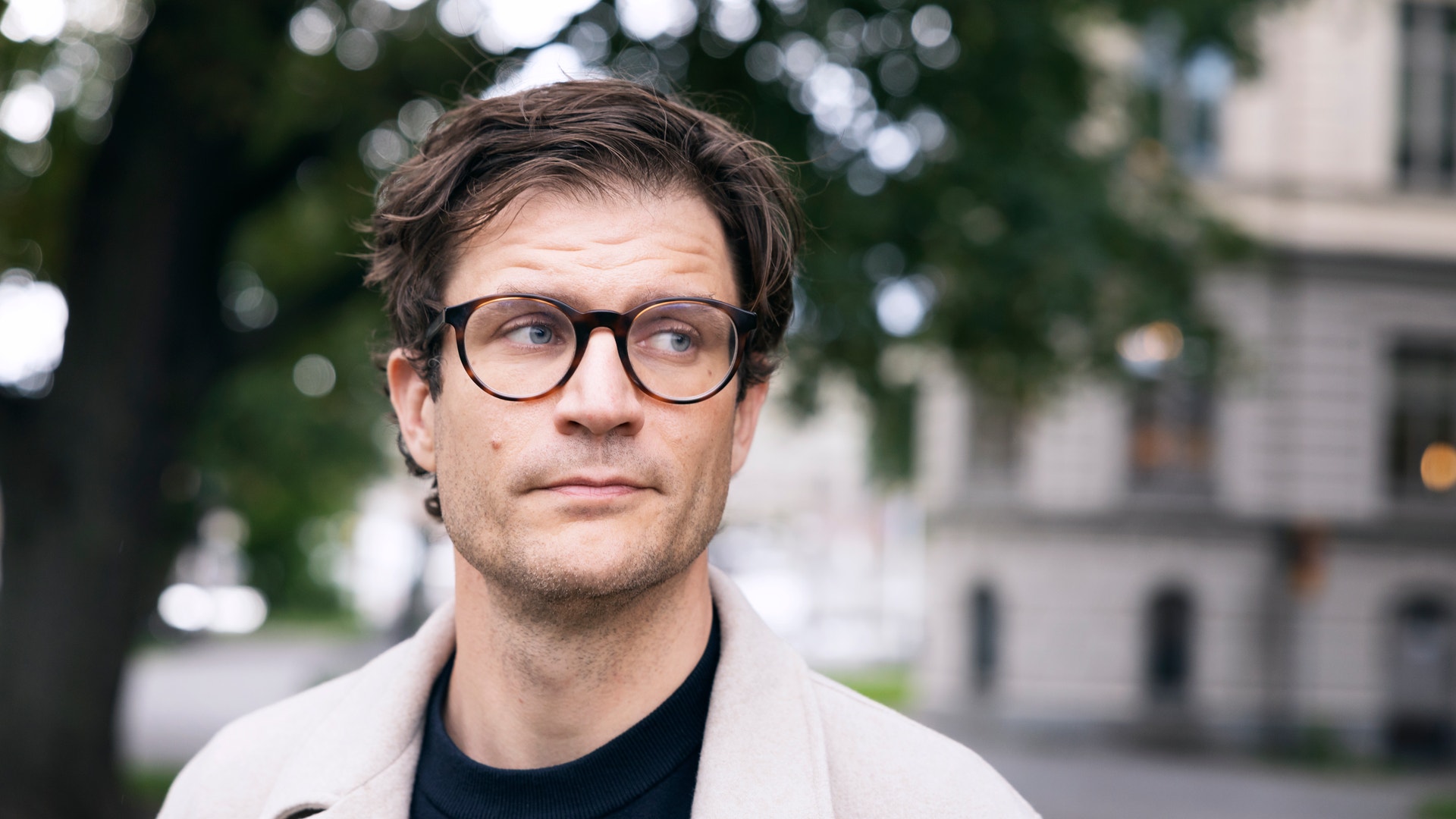 Porträtt av en man med glasögon och ljus jacka som står utomhus framför ett byggnadsliknande objekt.