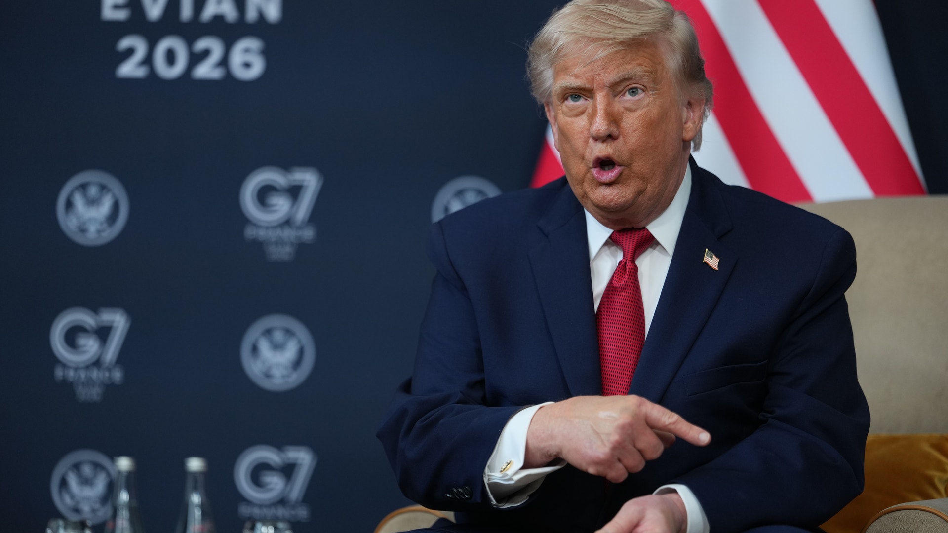 Man i mörkblå kostym och röd slips talar vid ett G7-möte i Evian 2026, med den amerikanska flaggan i bakgrunden.