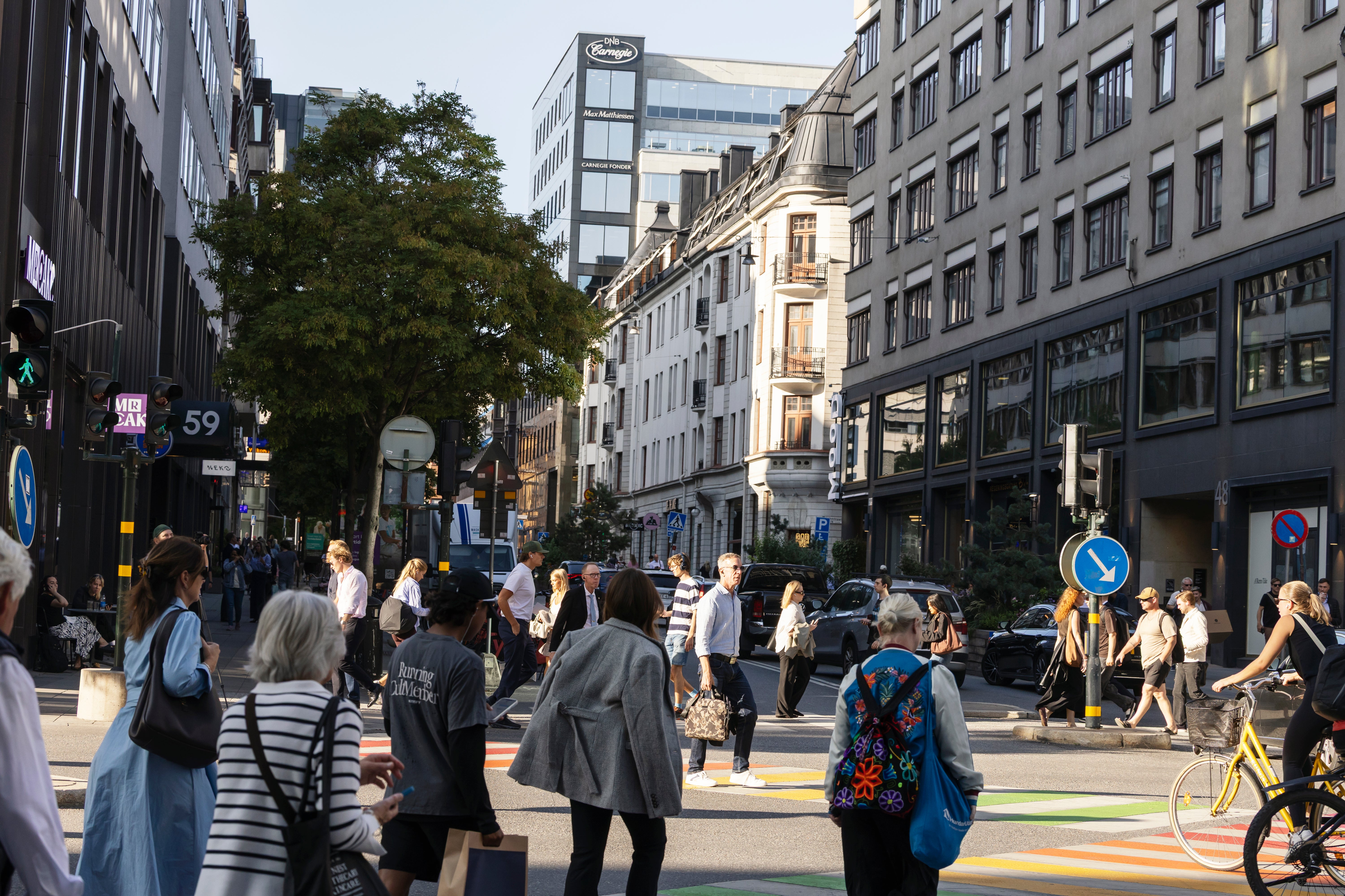 En livlig stadsgata med många människor som går omkring mellan klassiska och moderna byggnader. Människor i olika kläder går på en bred gata med cykelväg, omgivna av butiker och träd.